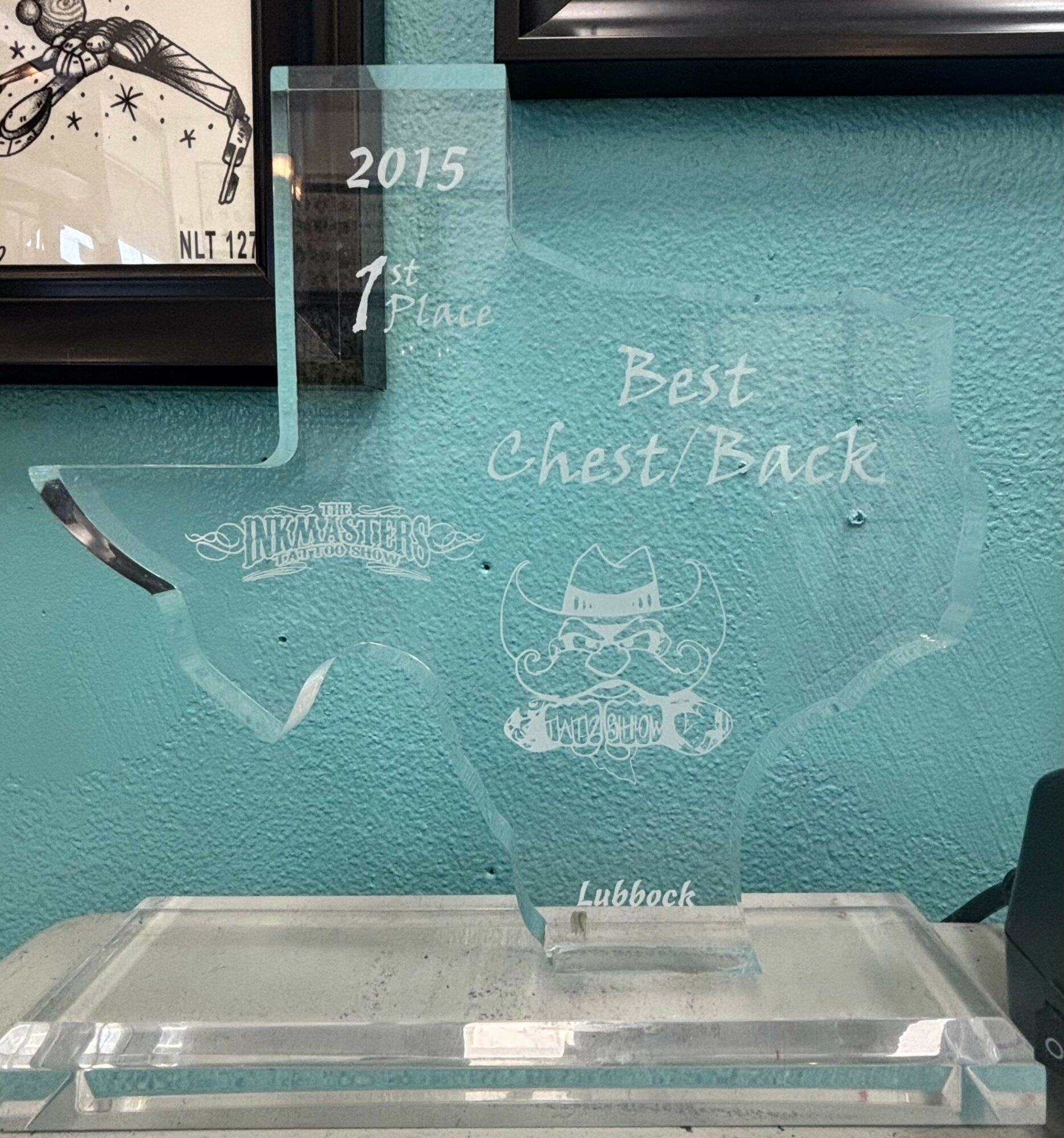 Clear glass trophy with engraved text and a cartoonish owl, placed on a table against a teal wall with framed pictures.