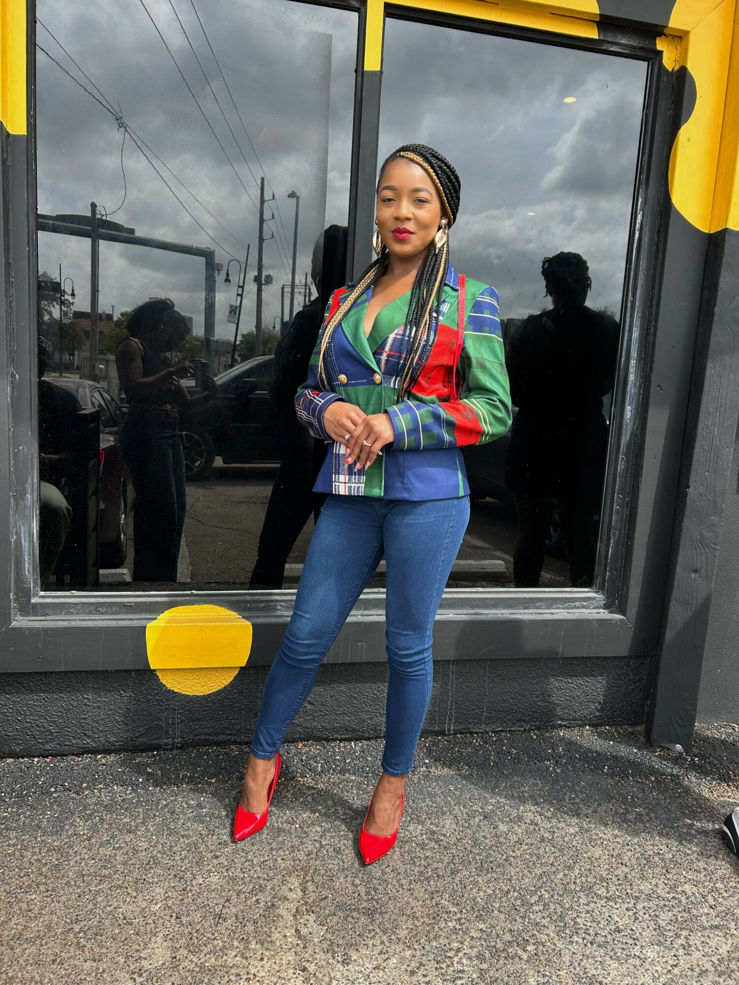 Woman standing outside a window, wearing a colorful jacket, blue jeans, and red high heels, smiling at the camera.