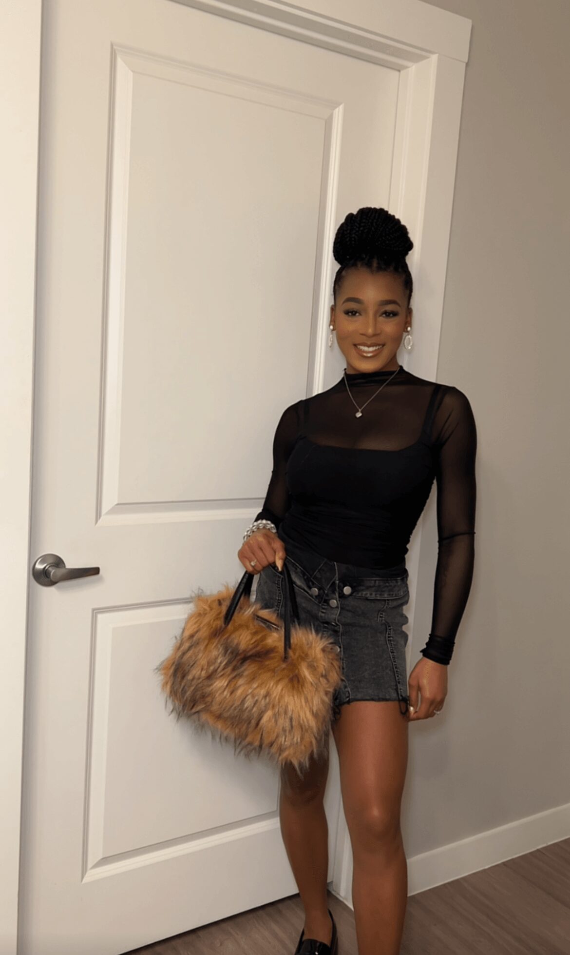 Young woman with dark hair in a bun, wearing a black sheer top and denim shorts, holding a furry handbag, smiling.