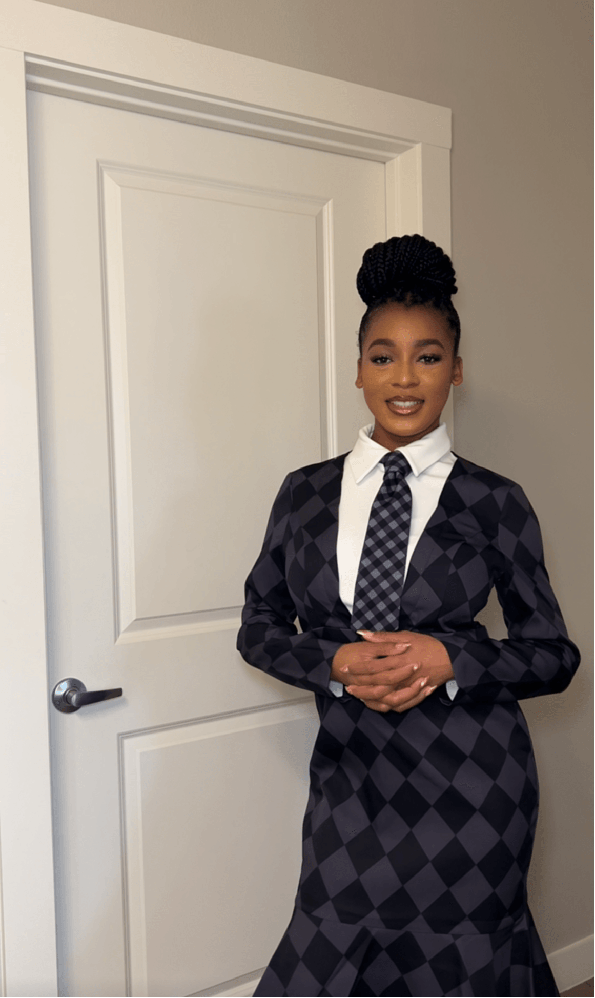Young woman in formal attire standing in front of a white door, smiling, with hands clasped.