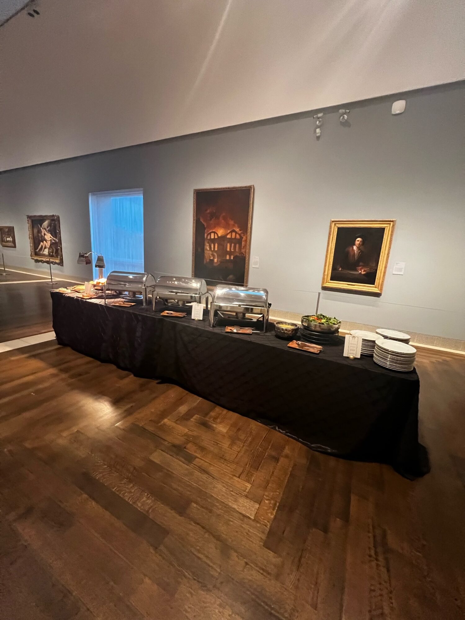 Buffet table with food containers and plates in front of a wall with framed artwork, hardwood floor, and a window.