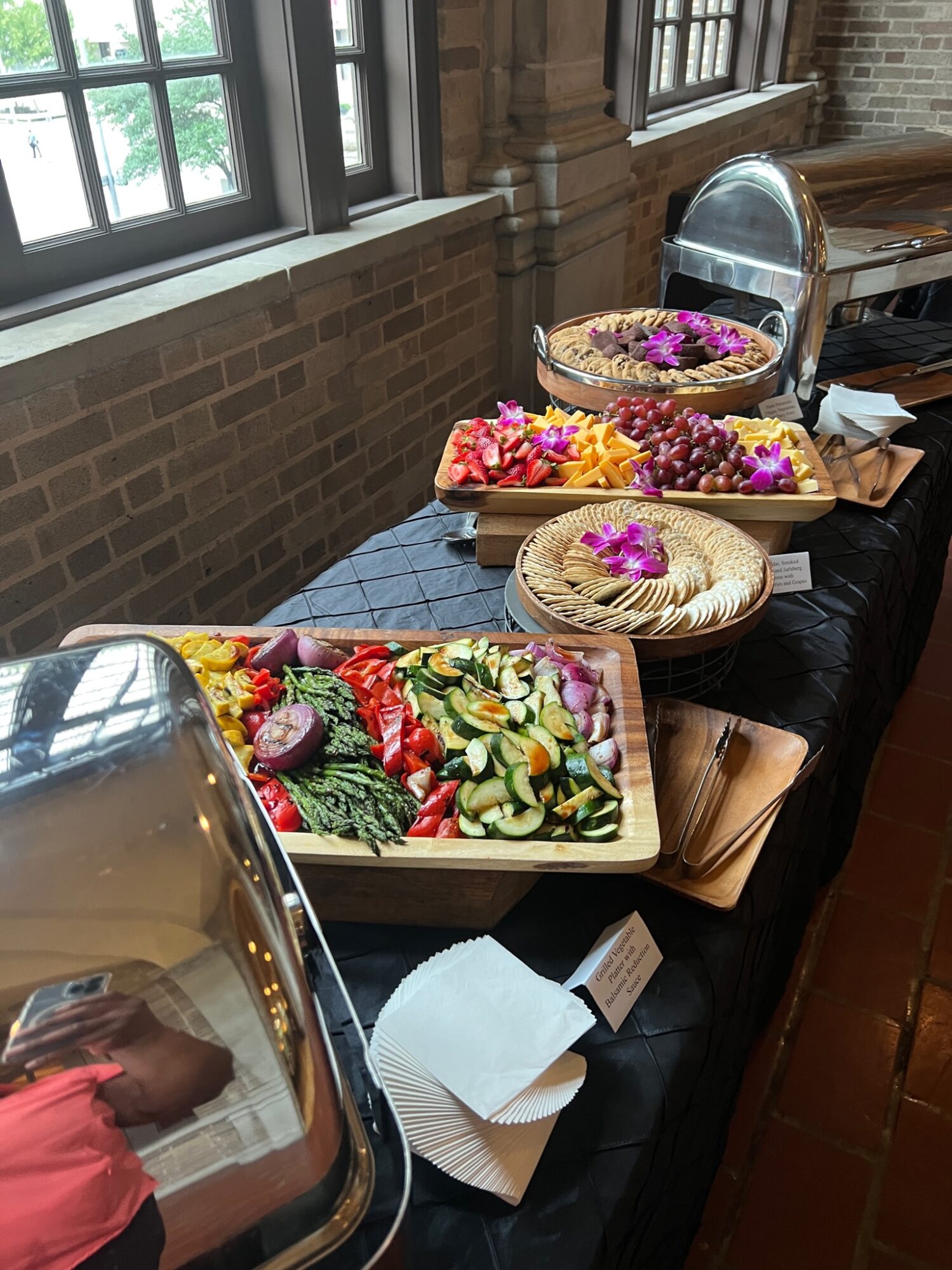 Display of colorful salads and dishes on a table near windows with brick walls, with serving trays and flowers.