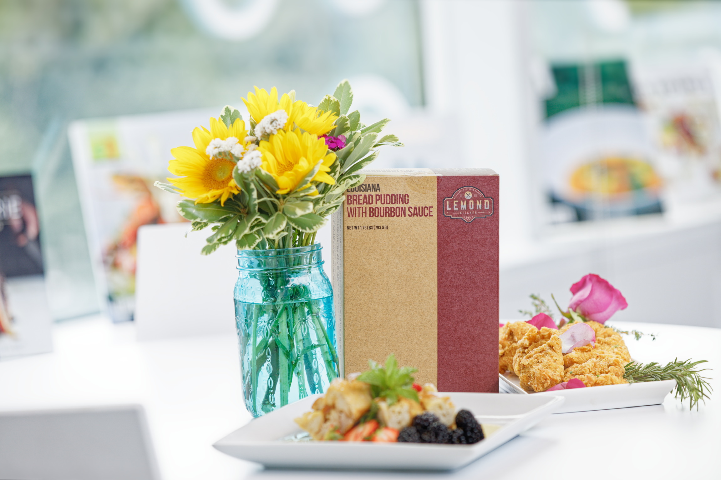 Vase with yellow sunflowers and white flowers on a table, with food plates and a menu in the background.