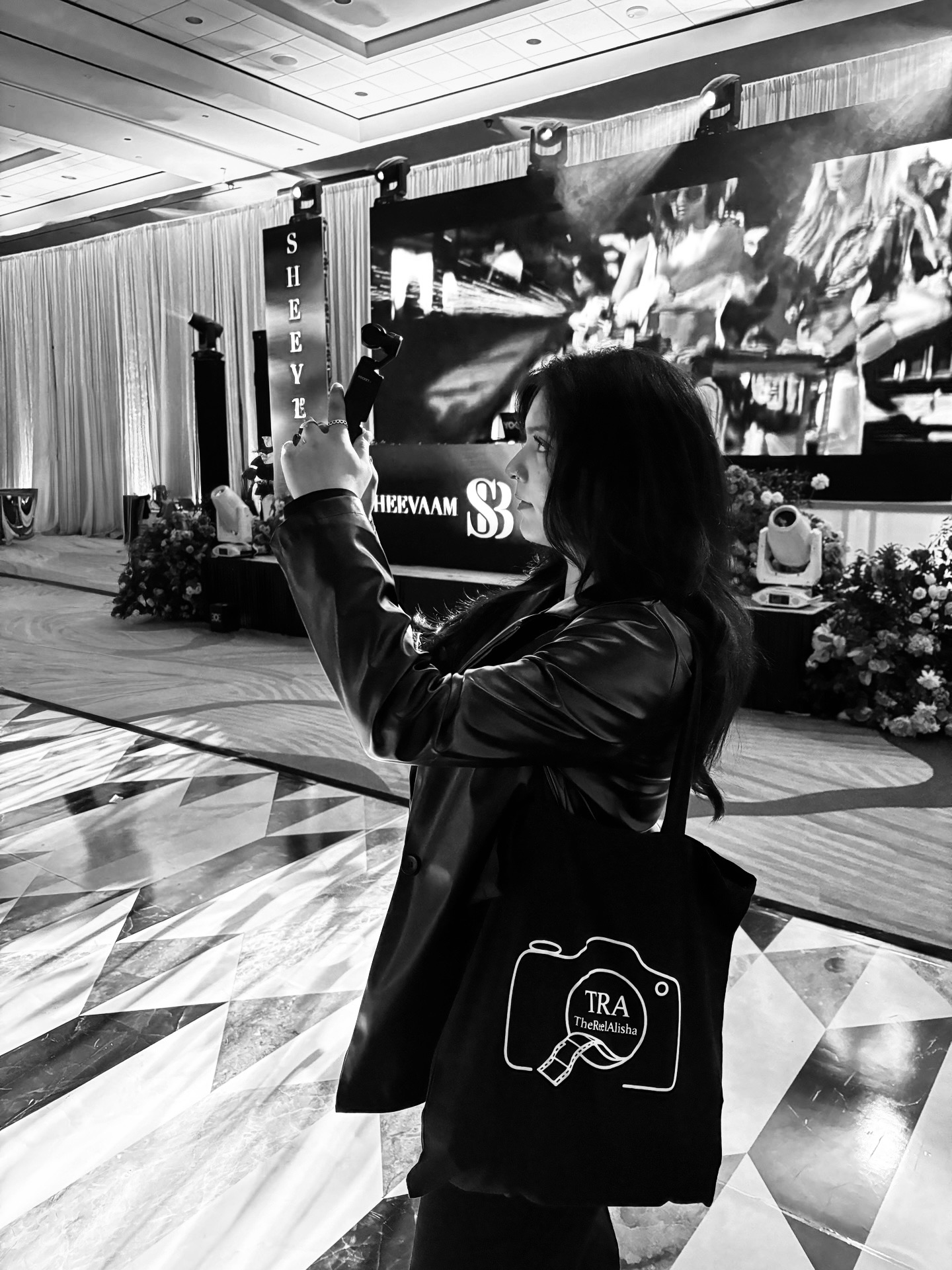 Person taking a photo with a smartphone at an indoor event with a large screen and stage in the background.