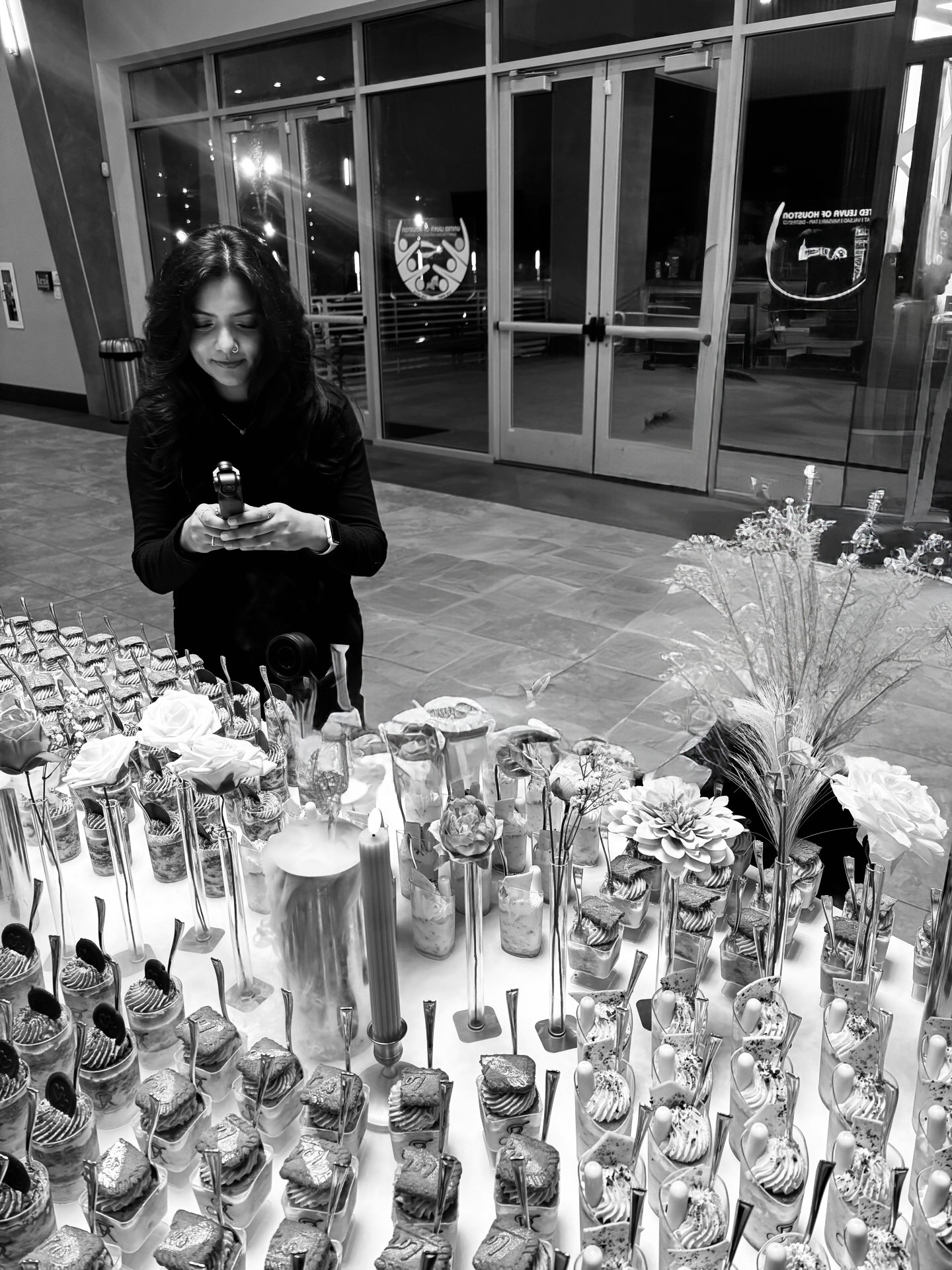 Woman with long hair looking at her phone in front of a table with various bottles and flowers, glass doors behind her.