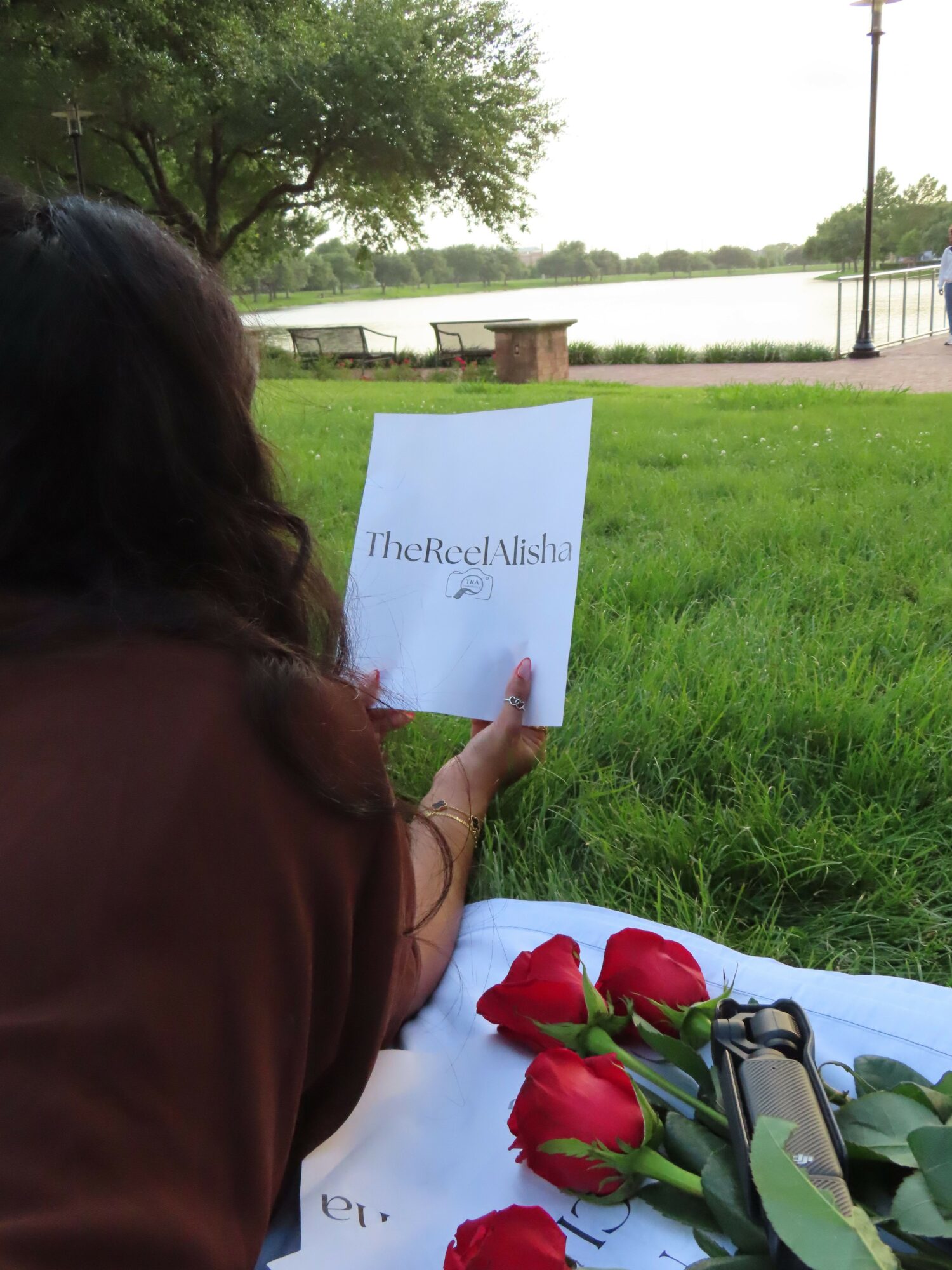 Person holding a sign with text, standing outdoors near water and trees, with a table of red roses nearby.