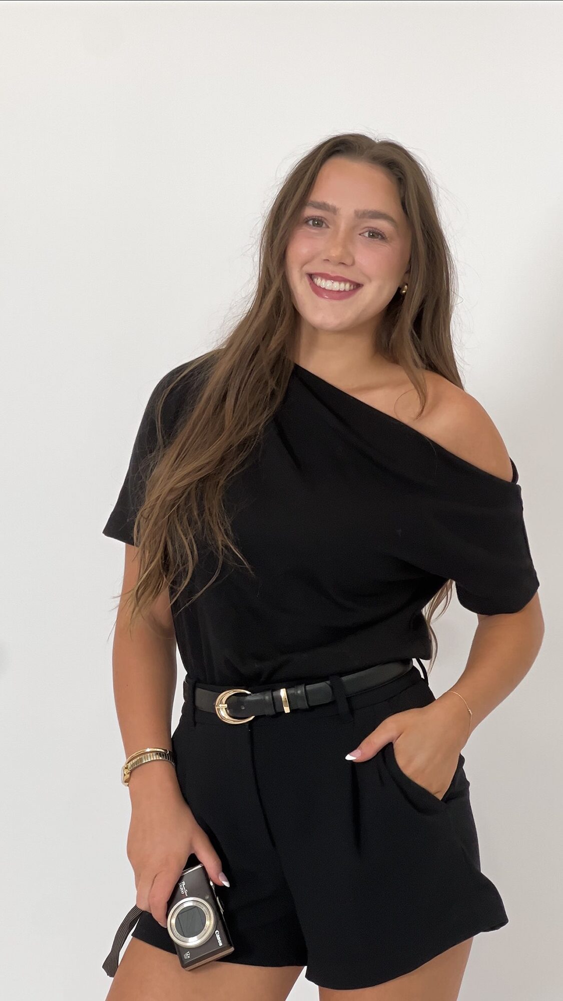 Young woman with long hair smiling, wearing a black off-shoulder top and black shorts, holding a camera, standing against a white background.