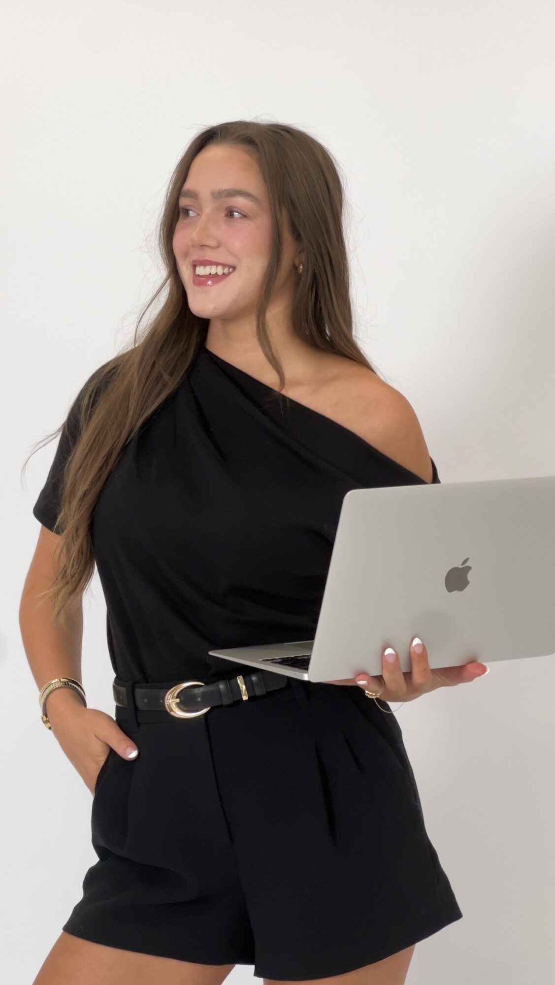 Young woman with long brown hair smiling, holding a silver laptop, wearing a black off-shoulder top and shorts, standing against a white background.