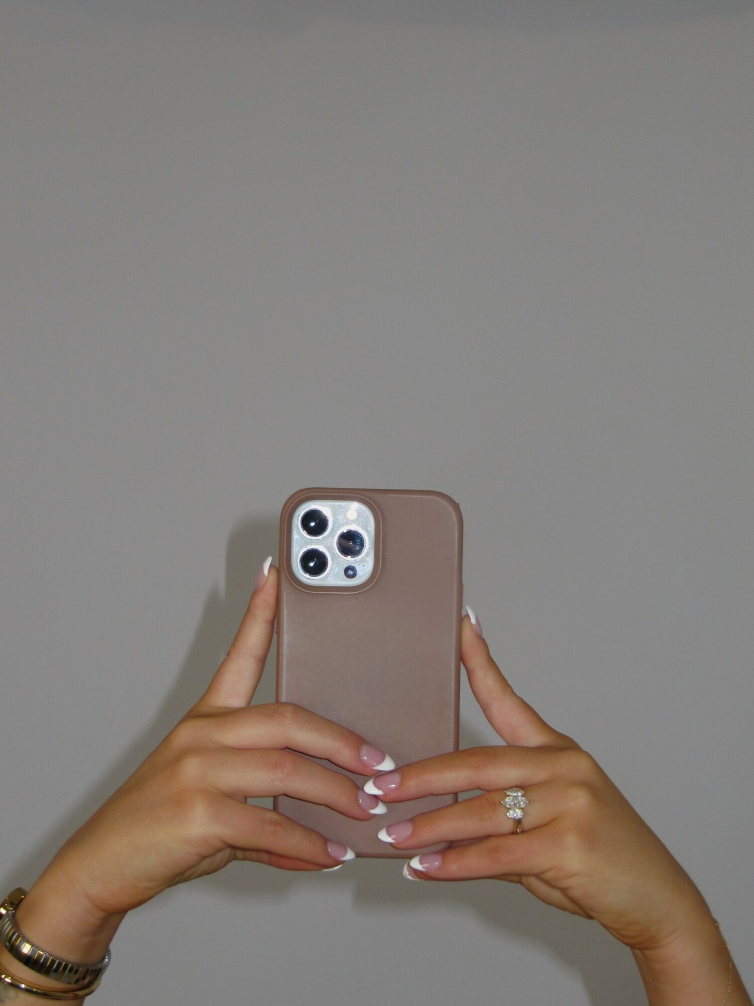 Person holding a smartphone with a brown case, showing hands with rings and a bracelet, against a plain gray background.