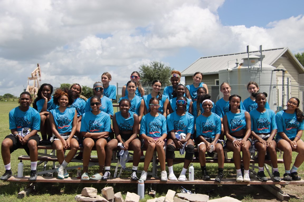 Group of young girls in matching blue shirts sitting on a train track outdoors, with a house and cloudy sky in background.