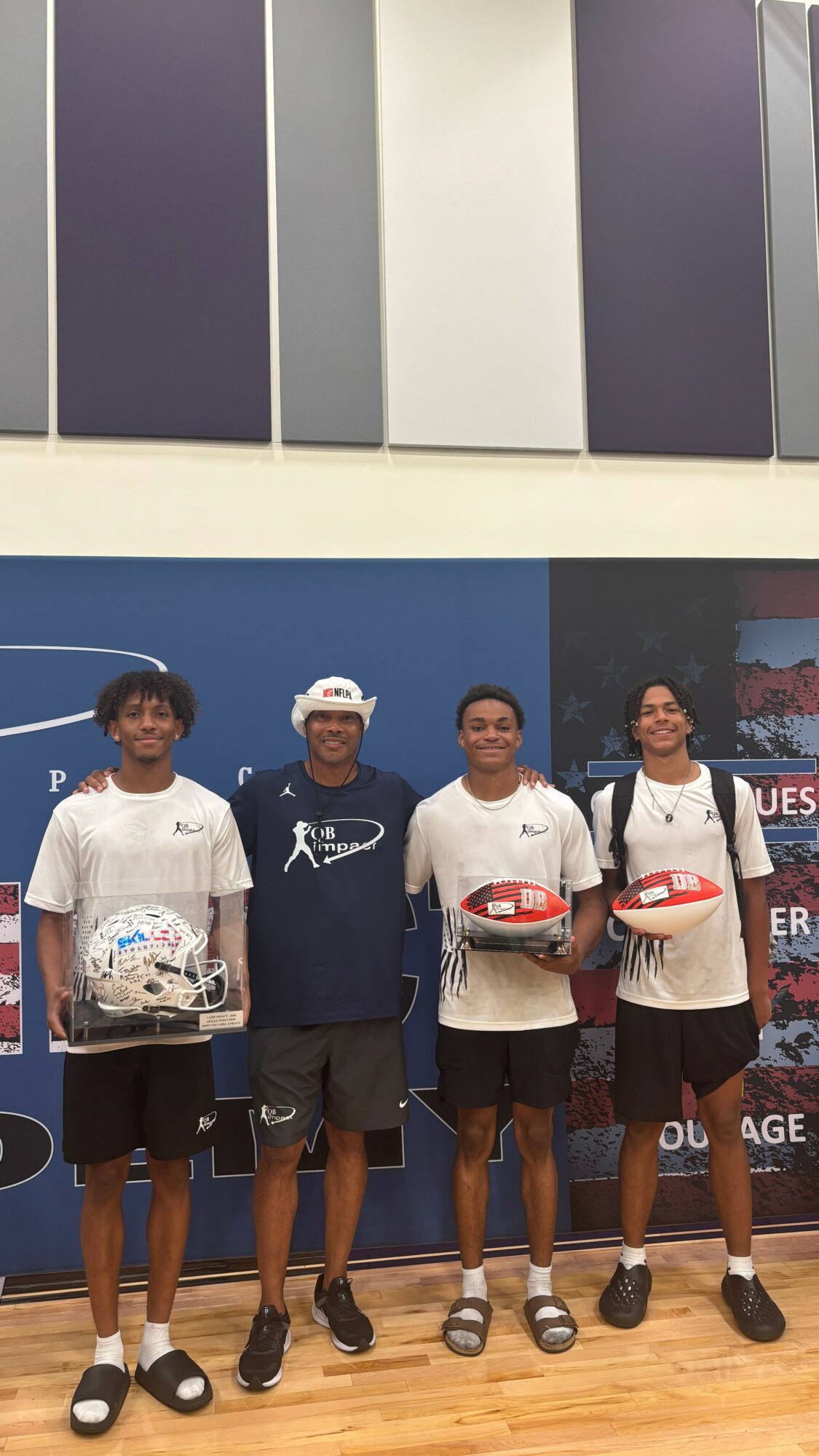 Four people standing indoors, holding helmets, smiling, with a blue wall and posters behind them.