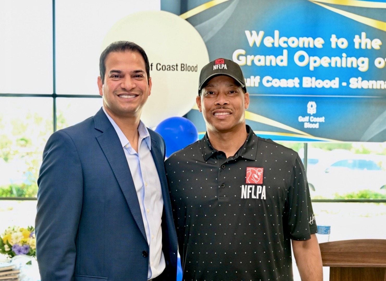Two men standing indoors, smiling, with a banner in the background welcoming to a grand opening event.