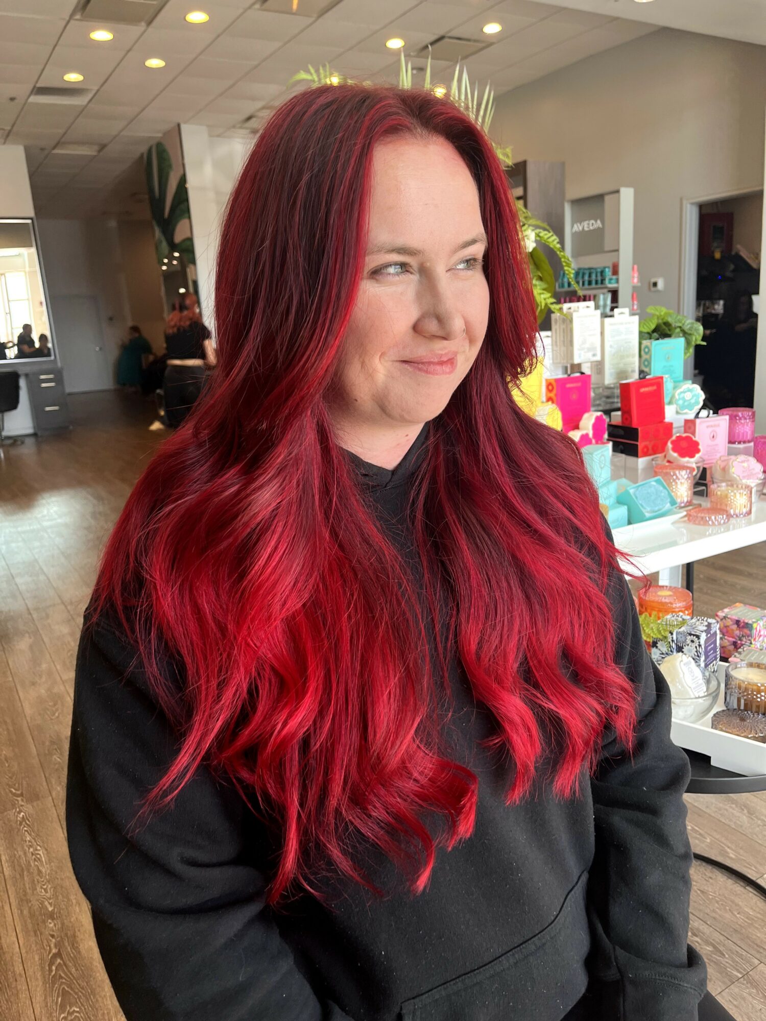 Woman with long, wavy red hair looking to the side in a store, with shelves of colorful products behind her.
