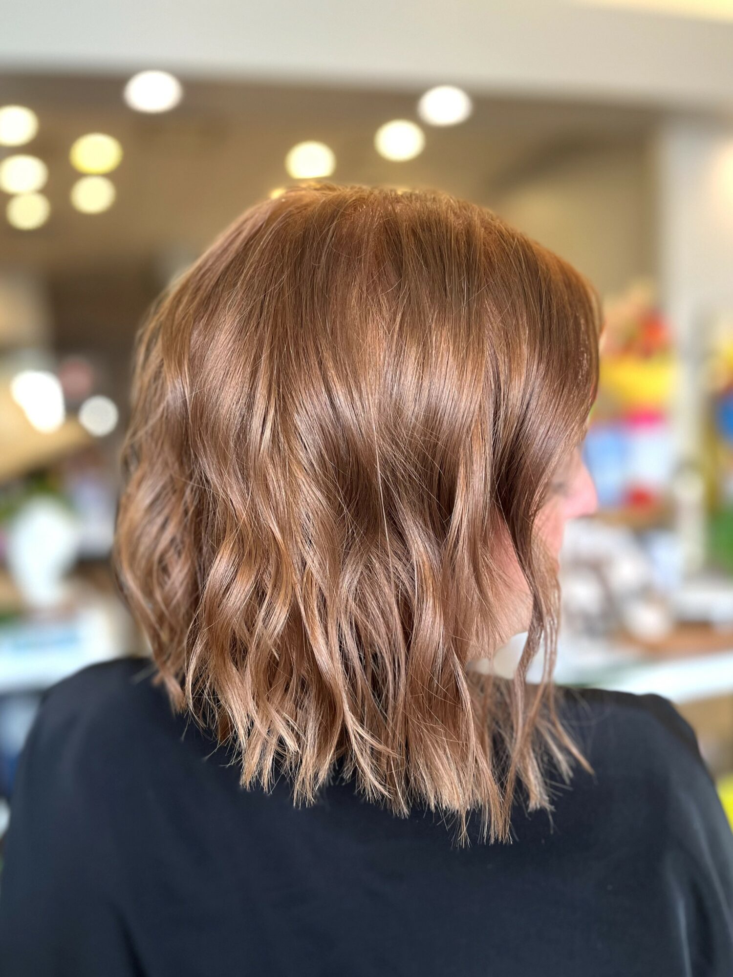Back of a person's head with shoulder-length wavy brown hair, wearing a dark top, in an indoor setting.