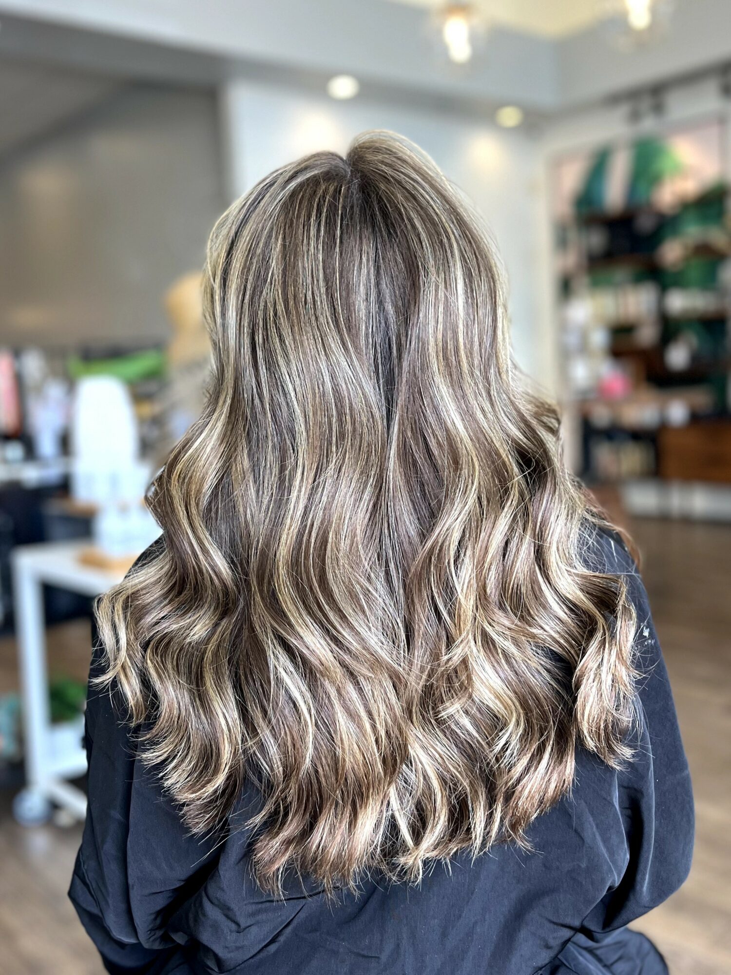 Back view of long, wavy blonde hair in a salon setting with shelves and equipment visible