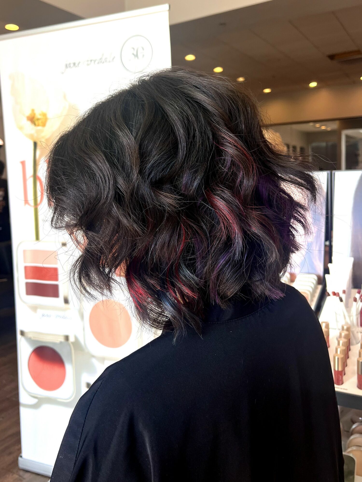 Side view of a woman with wavy, dark hair with purple highlights, wearing a black top, in a store setting.