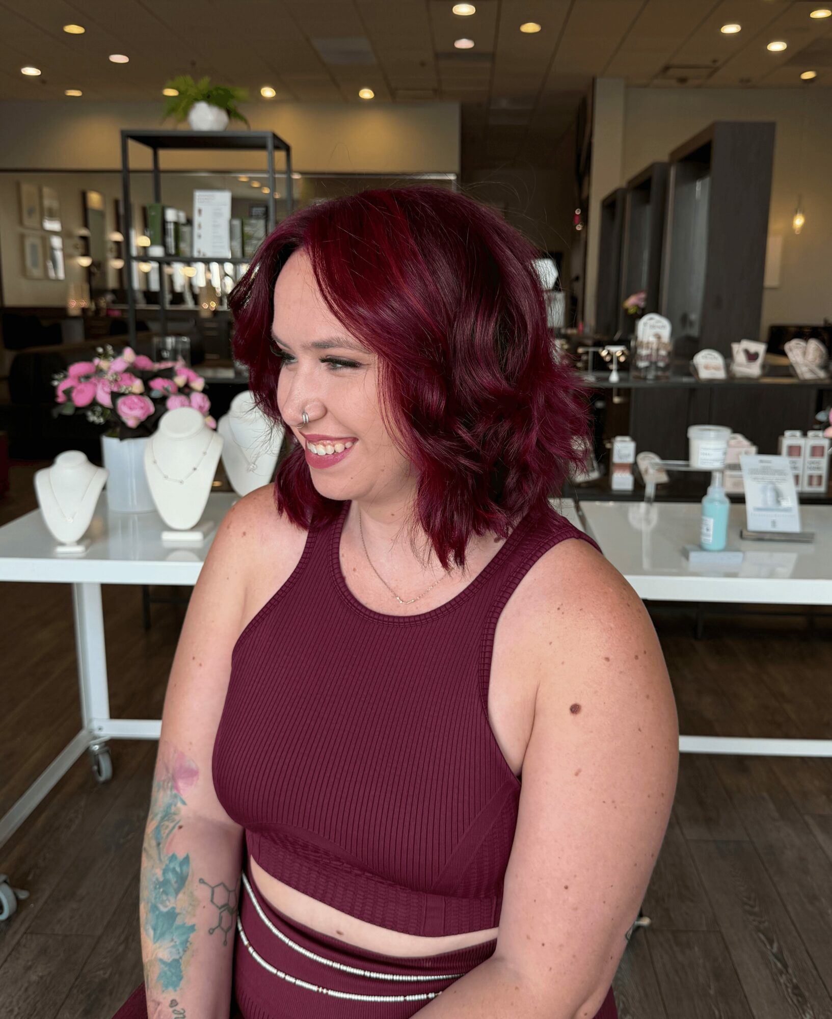 Woman with red hair smiling, wearing a sleeveless maroon top, standing indoors with display tables and flowers in background.