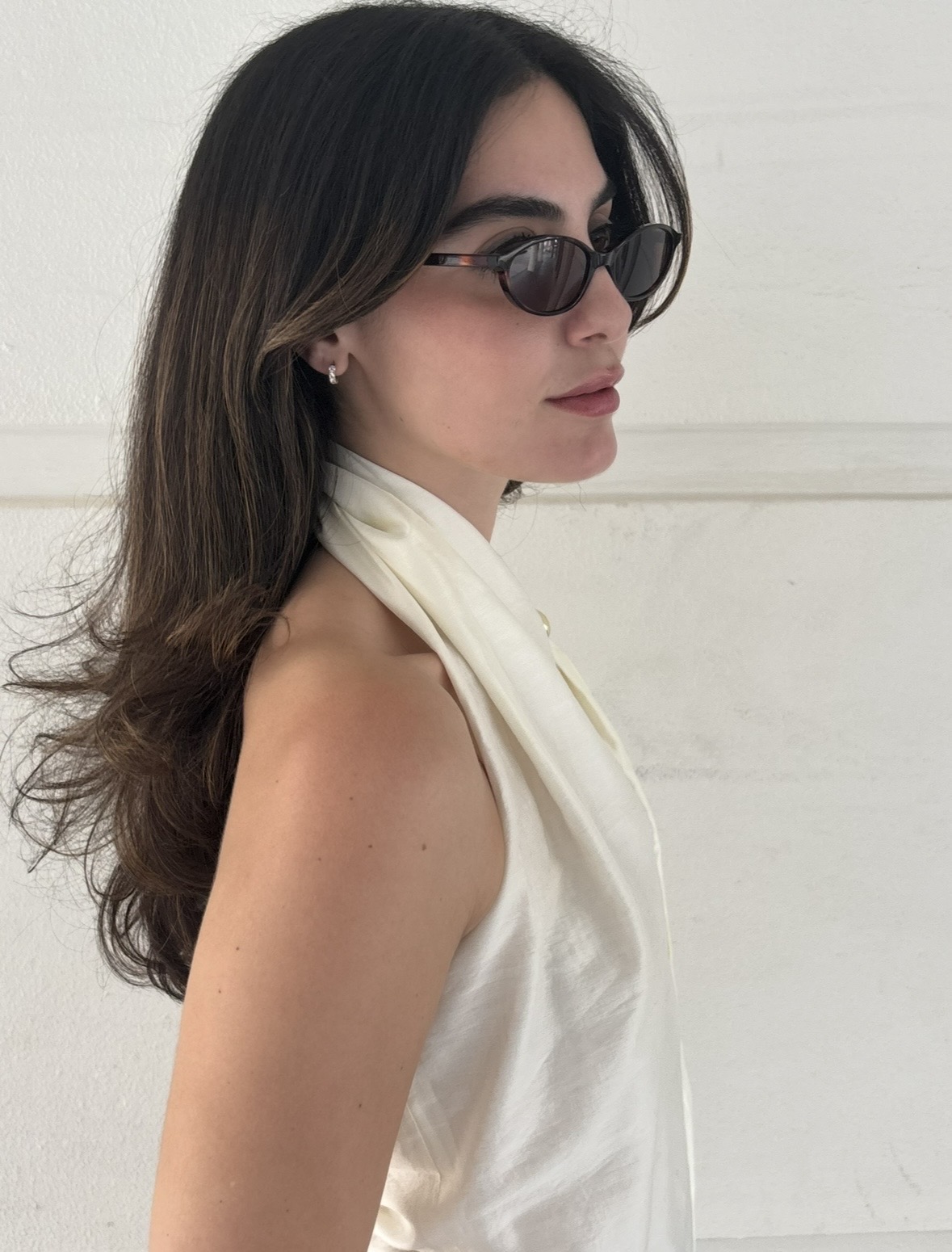 Woman with long dark hair wearing sunglasses and a white sleeveless top, facing sideways against a white background.