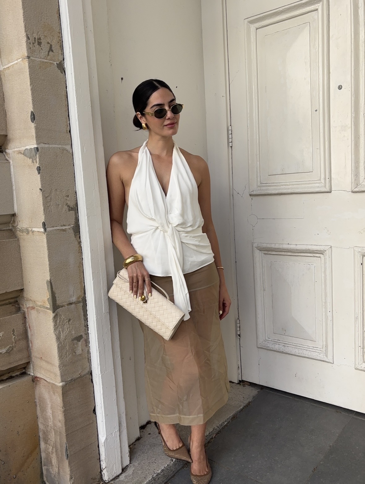 Woman in sunglasses and white top stands near a door, holding a beige clutch, wearing a beige skirt and heels.