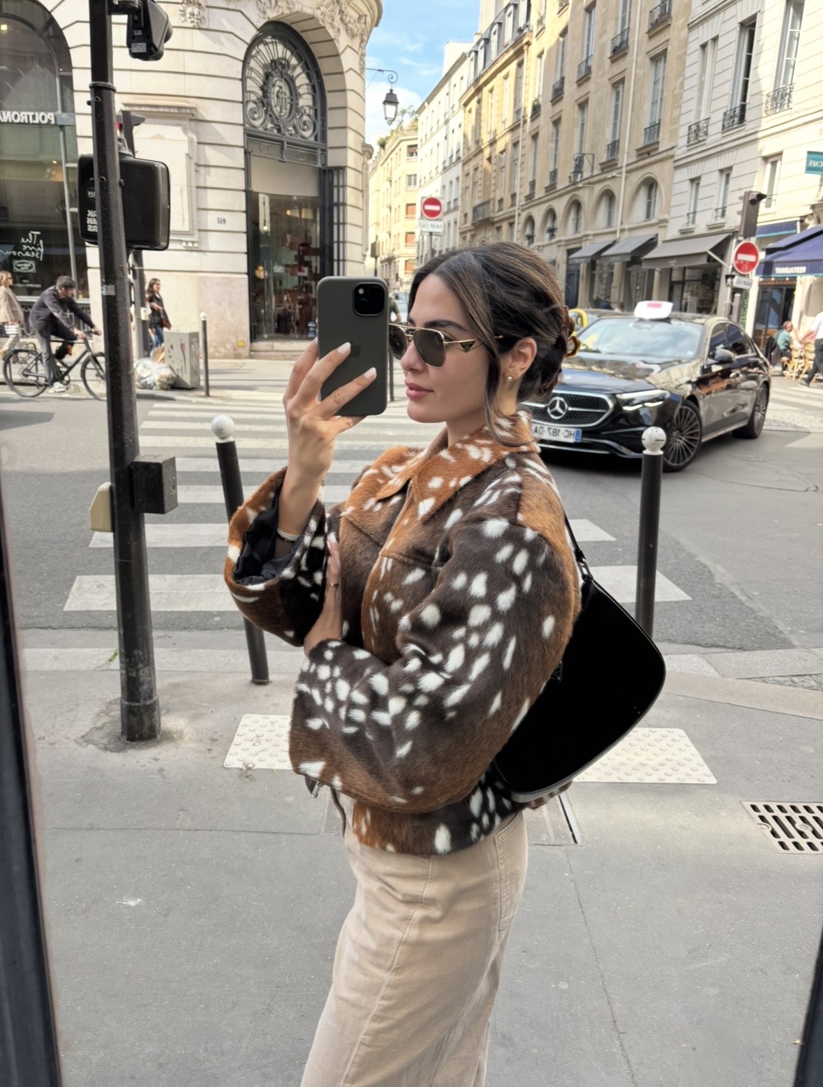 Woman taking a selfie on a city street, wearing sunglasses, a patterned jacket, and beige pants, with buildings and cars in background.