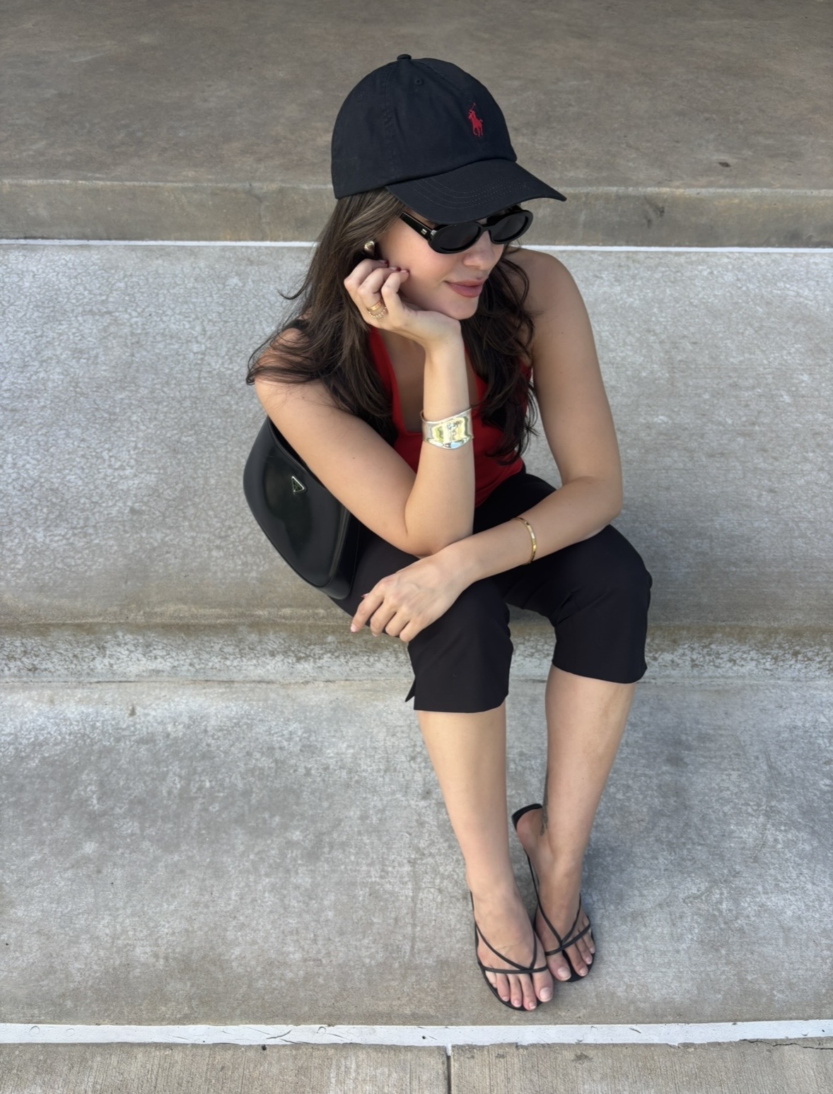 Young woman sitting on concrete steps, wearing a black cap, sunglasses, red top, black pants, and sandals, with a backpack.