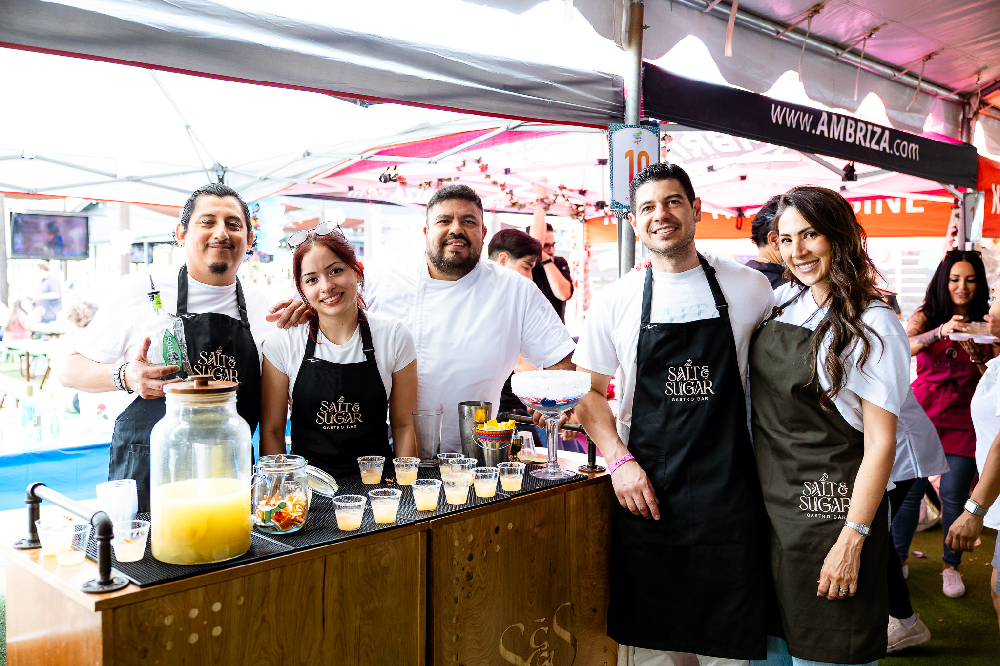Group of five people behind a food stall at an outdoor event, smiling and wearing aprons, with drinks and food items on the counter.