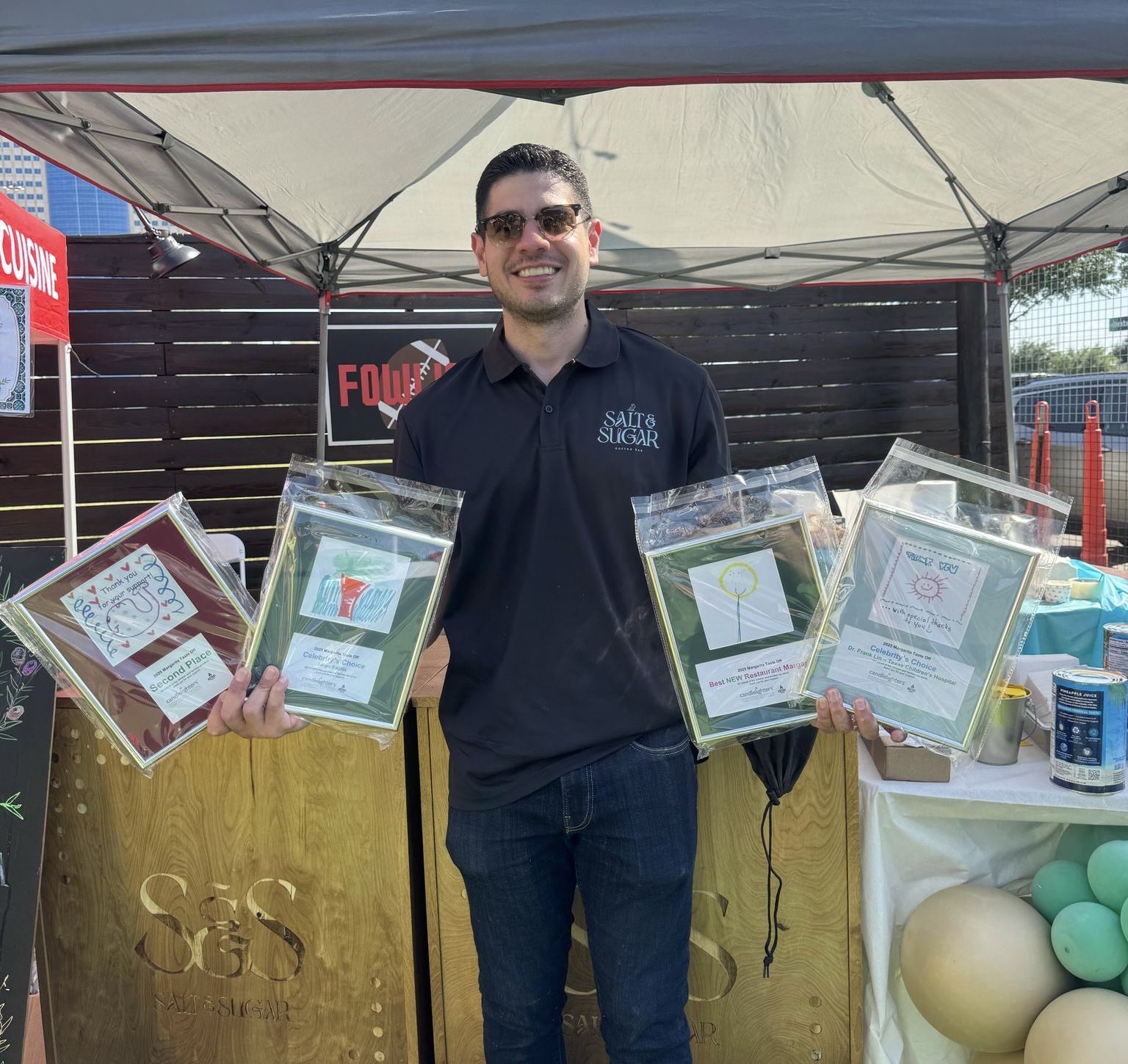 Man holding four framed certificates, smiling, standing under a canopy with balloons and a table behind him.