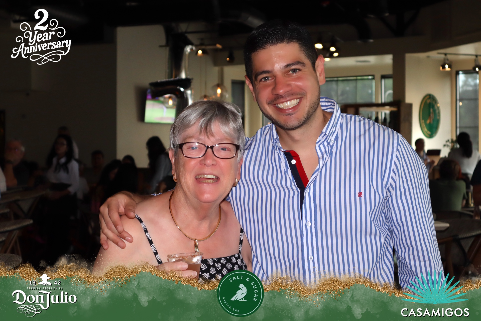 An elderly woman and a young man smile and pose together indoors, with a bar and people in the background.