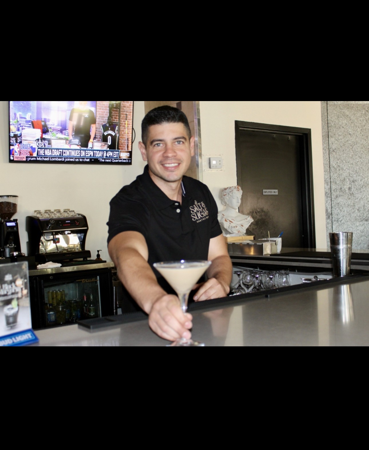 Smiling bartender holding a cocktail glass at a bar counter, with a TV and decor in the background.