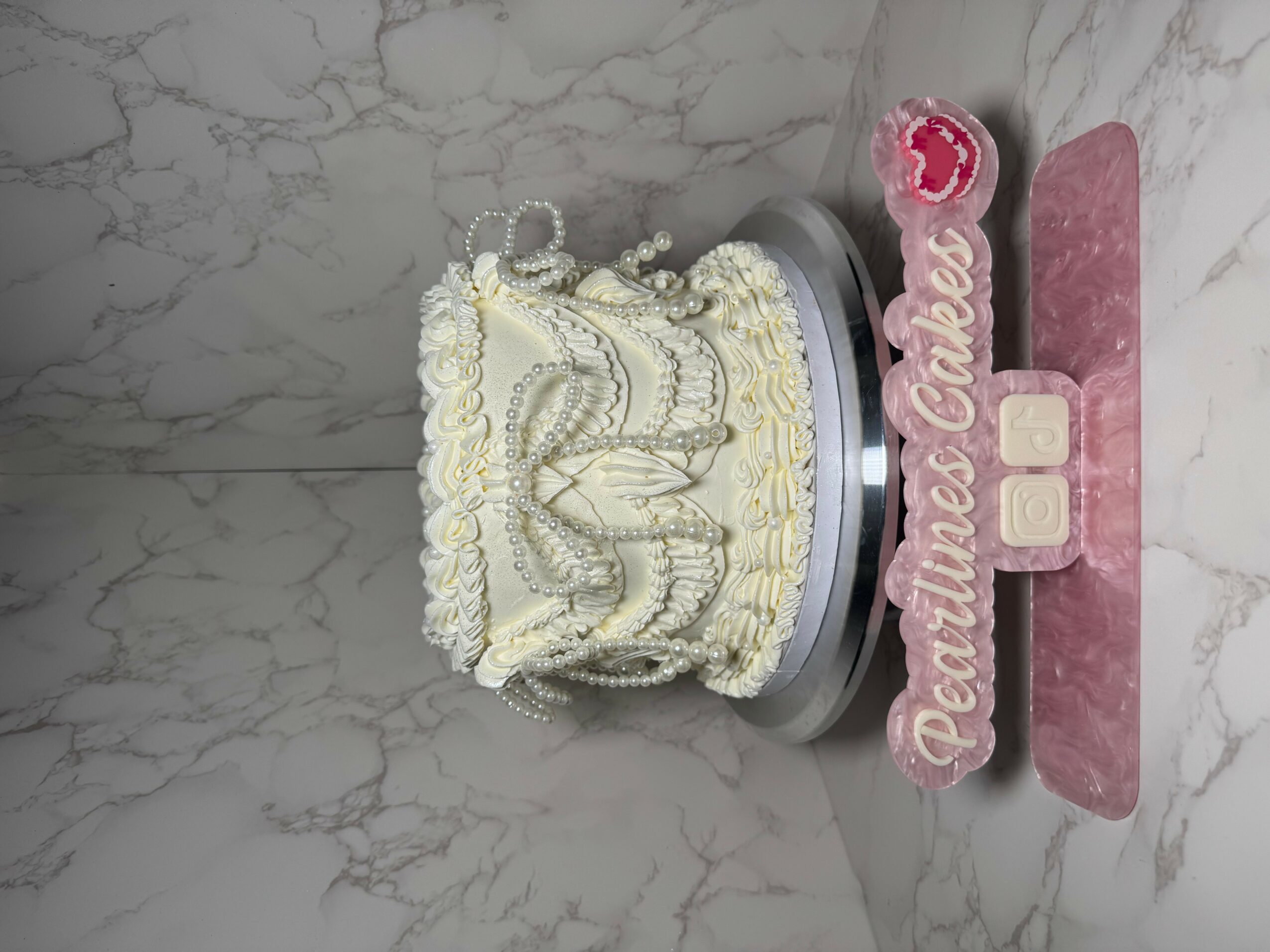 A decorated cake with white icing and pearl accents on a silver stand, with a pink sign reading 'Dreams Come True' in front.