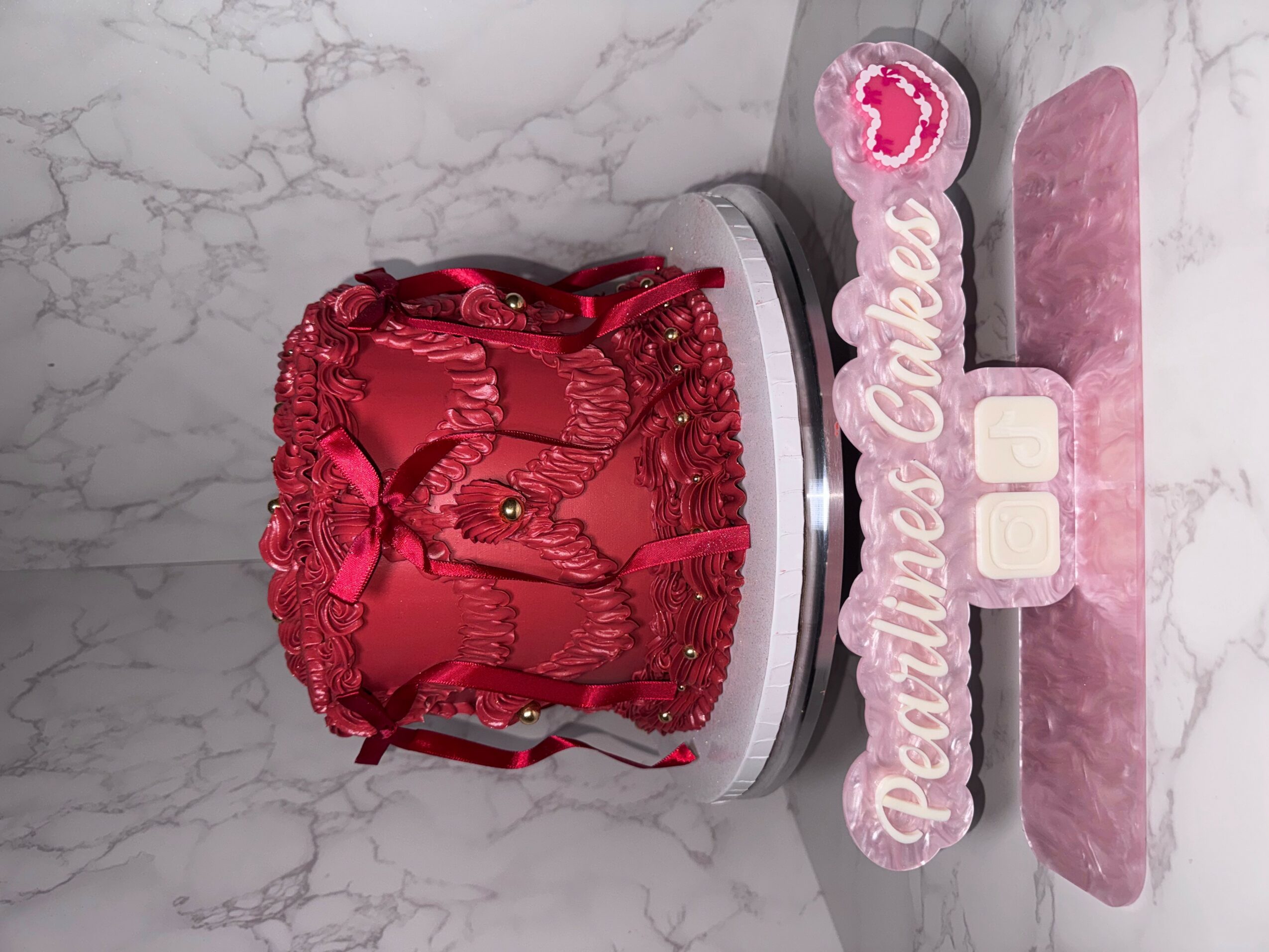 Pink and red decorative cake with a ribbon, on a pink and white base, with a pink sign reading 'Pettiness Queen' and two white blocks.