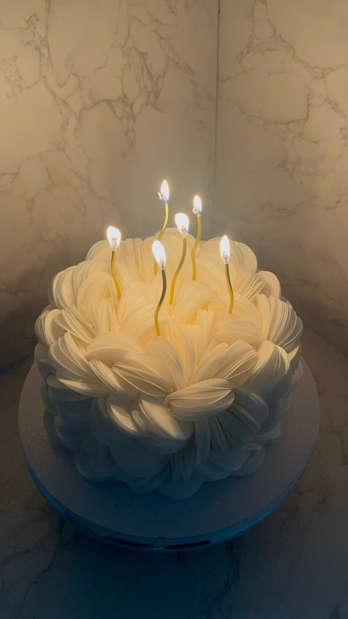 Round cake with white frosting and flower-like decorations, lit candles on top, on a marble surface.