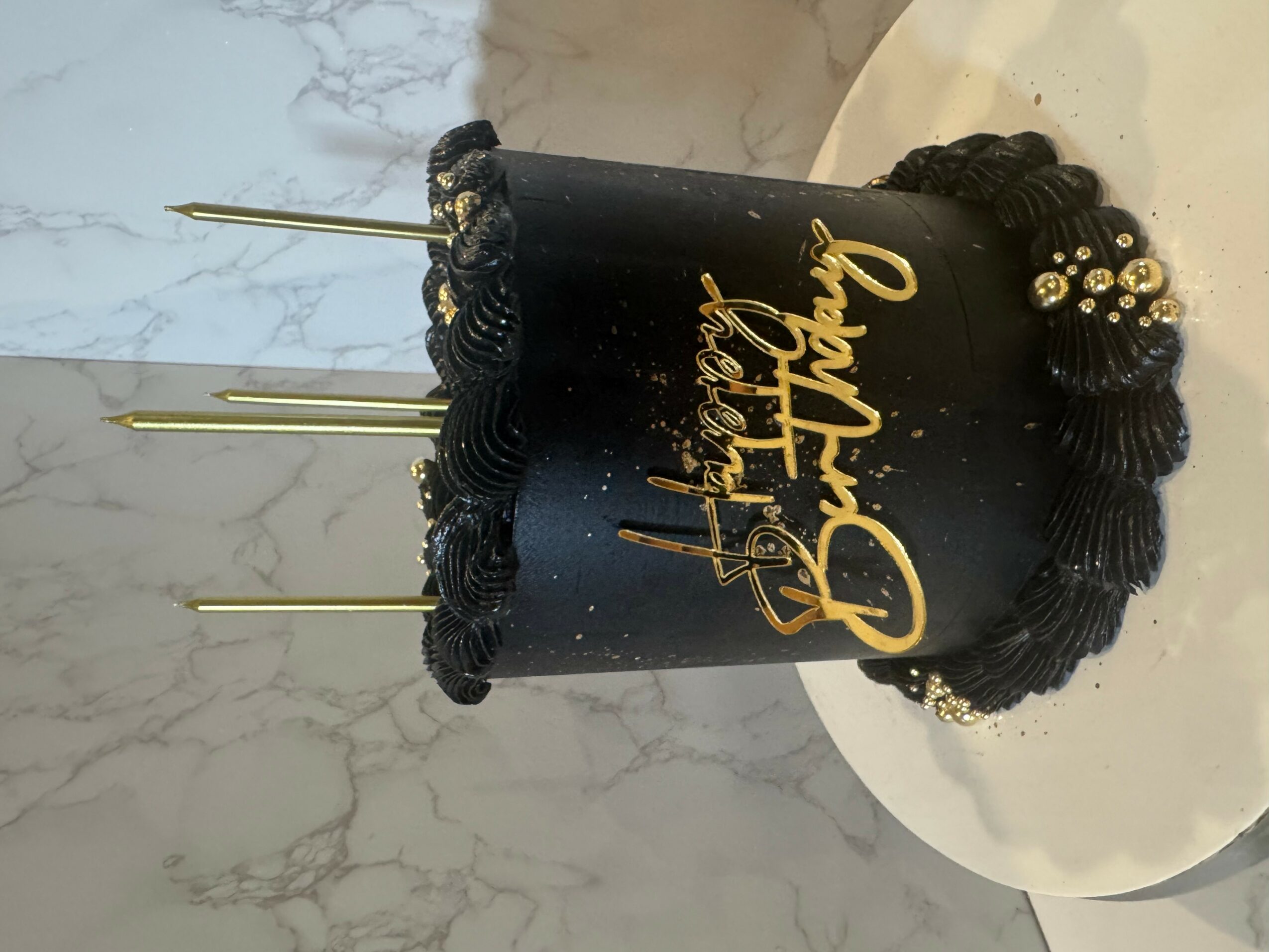 Black birthday cake with gold lettering and black decorations, three gold candles, on a white plate, marble background.