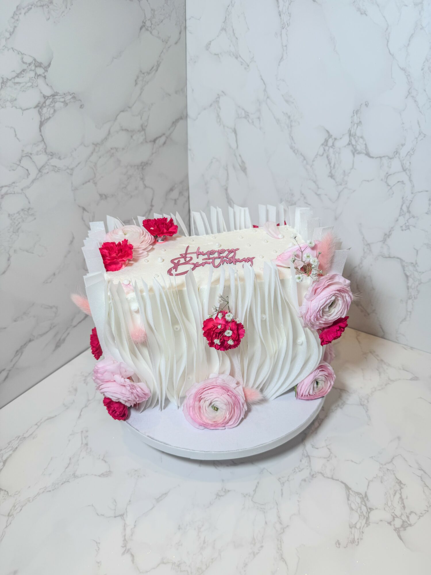 Decorative cake with pink and white flowers, pink meringues, and a pink message on top, set against a marble background.