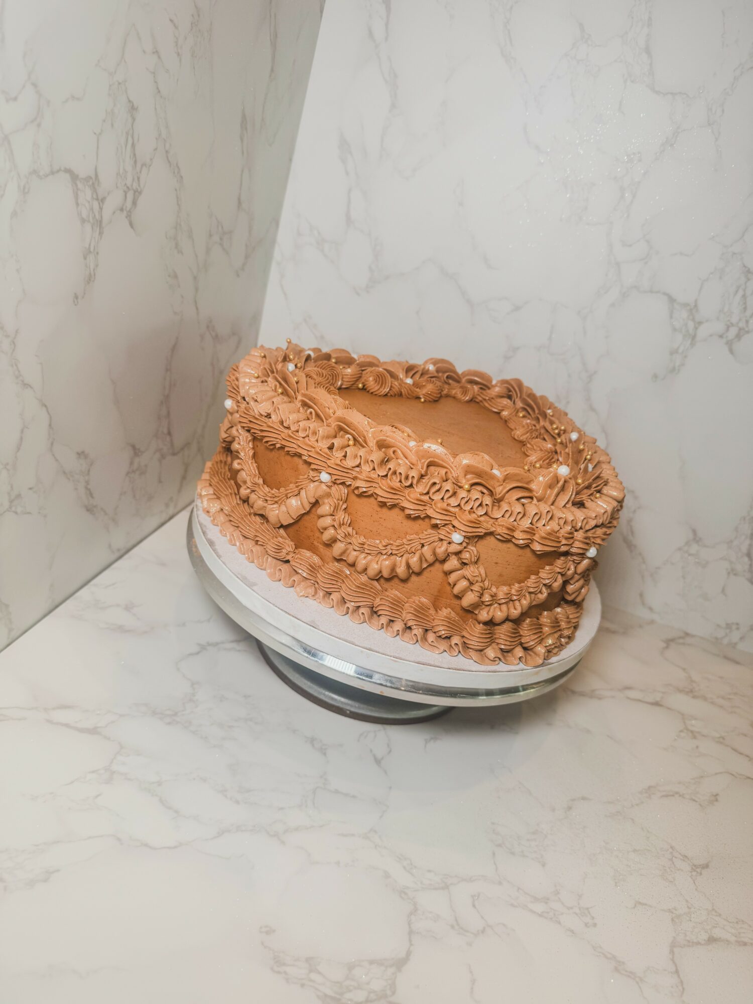 Round cake with peach-colored frosting and decorative piping, placed on a white marble surface against a marble wall background.