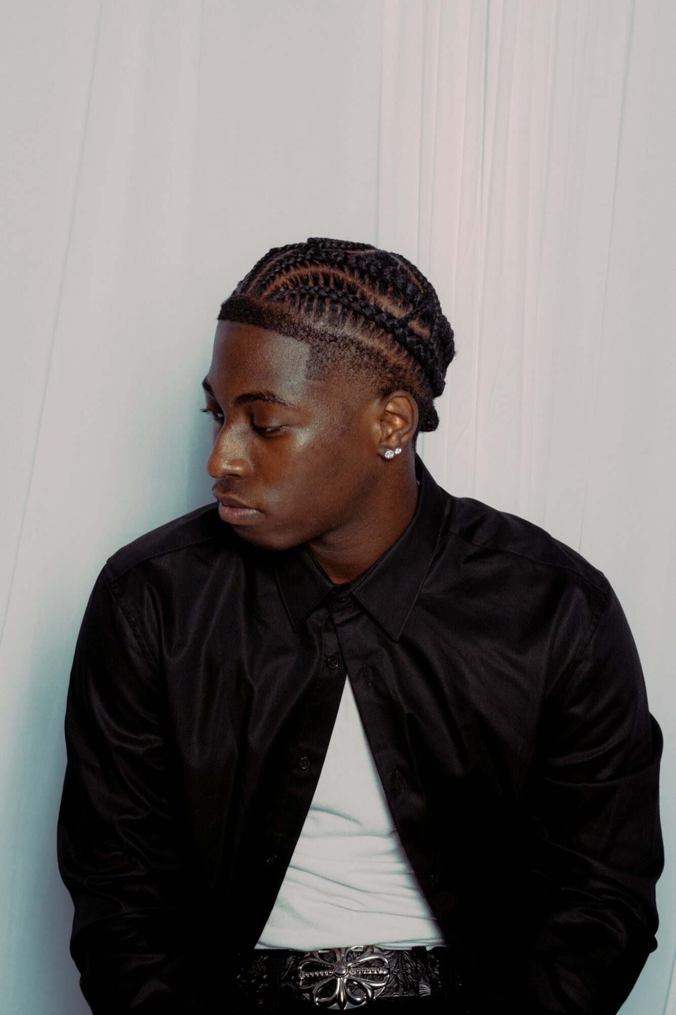 Side profile of a person with braided hairstyle, wearing a black jacket and white shirt, against a plain background.