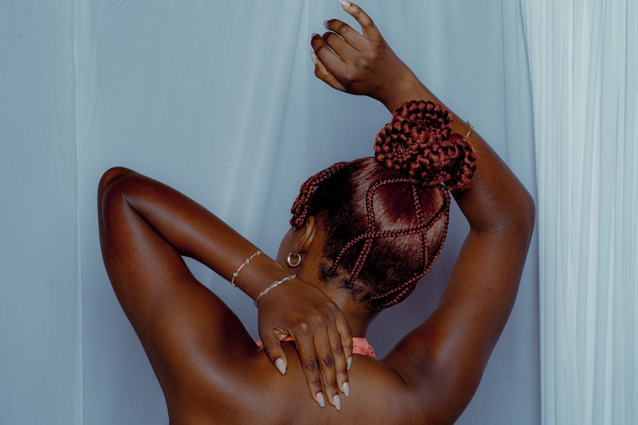 Person with braided hair and arm raised, covering face, against light blue curtain background.