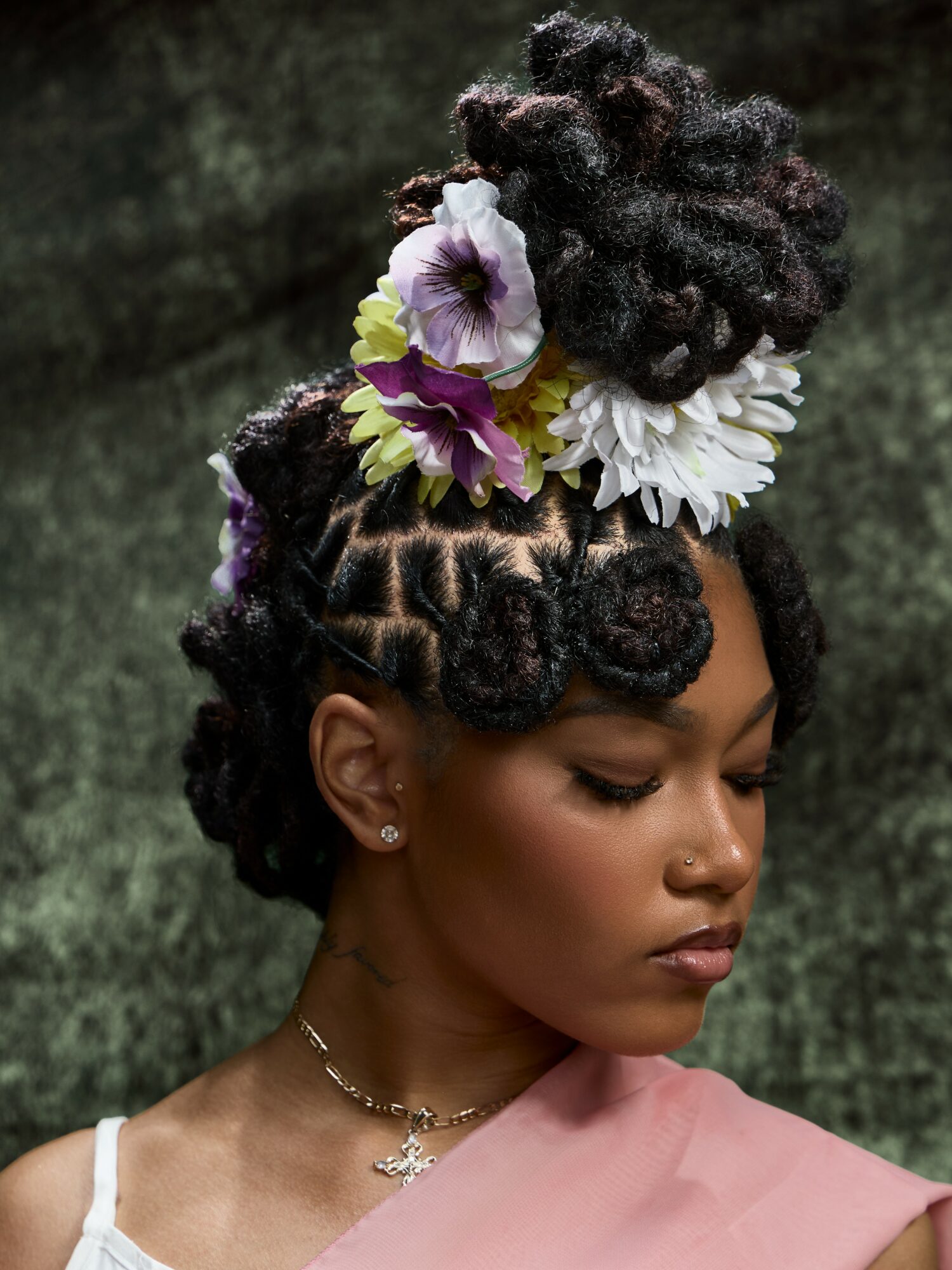 Young woman with styled hair adorned with large purple, white, and yellow flowers, wearing a pink top and necklace.