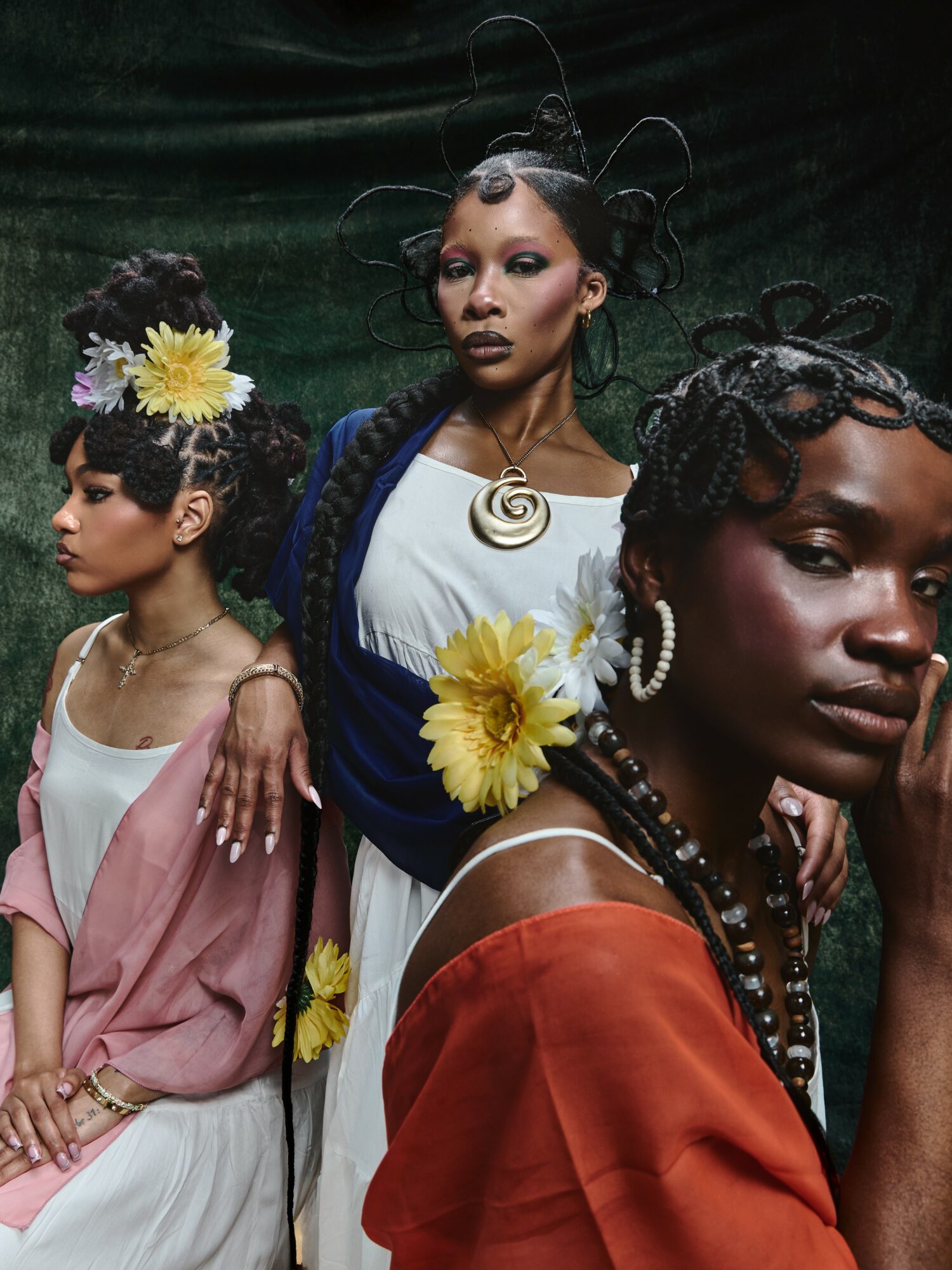 Three women with elaborate hairstyles and accessories pose together, wearing colorful clothing and floral decorations.