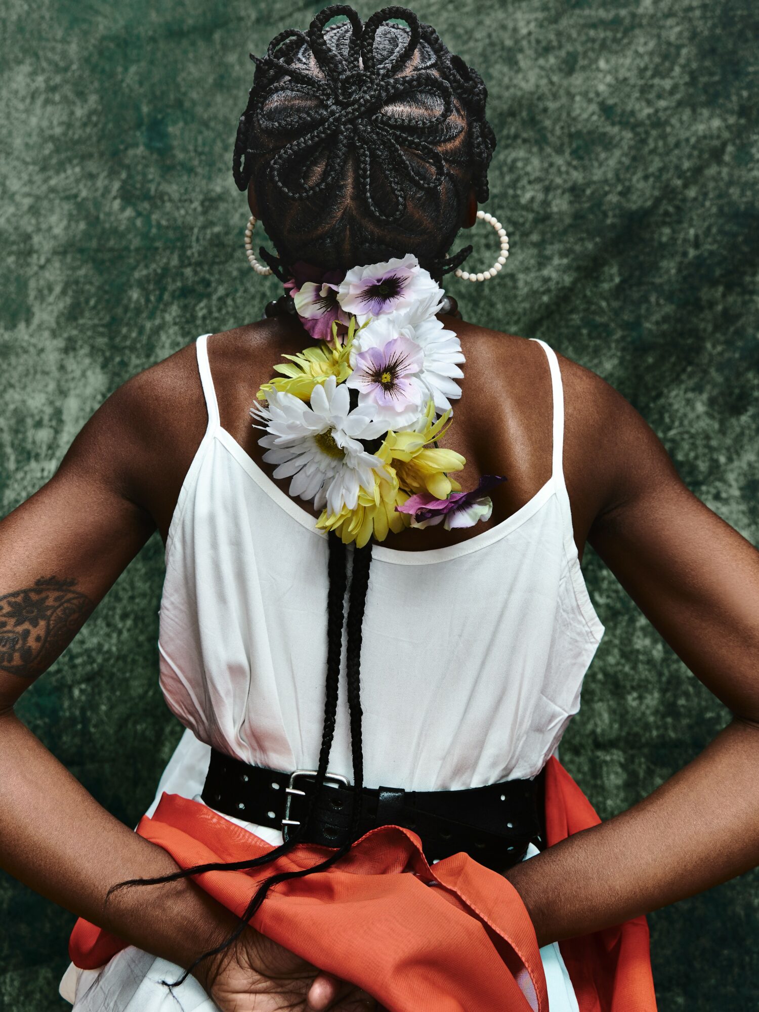 Back view of a woman with braided hair, wearing a white top, black belt, and holding an orange bag, against a green background.