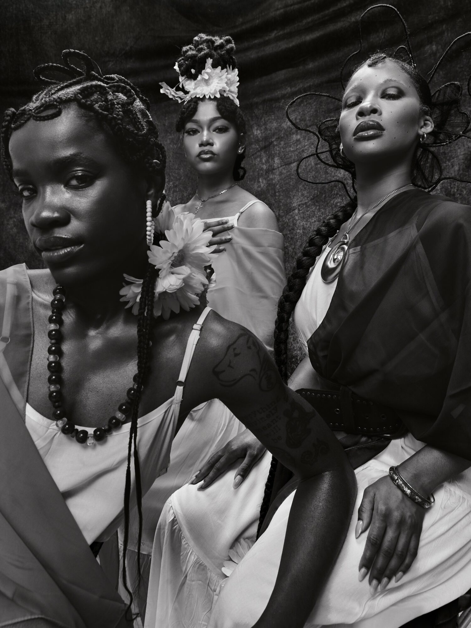 Three women with elaborate hairstyles and accessories pose against a dark background.