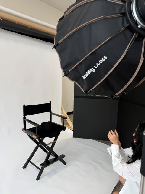 Studio setup with a large black umbrella light, a black director's chair, and a person in white clothing adjusting equipment.