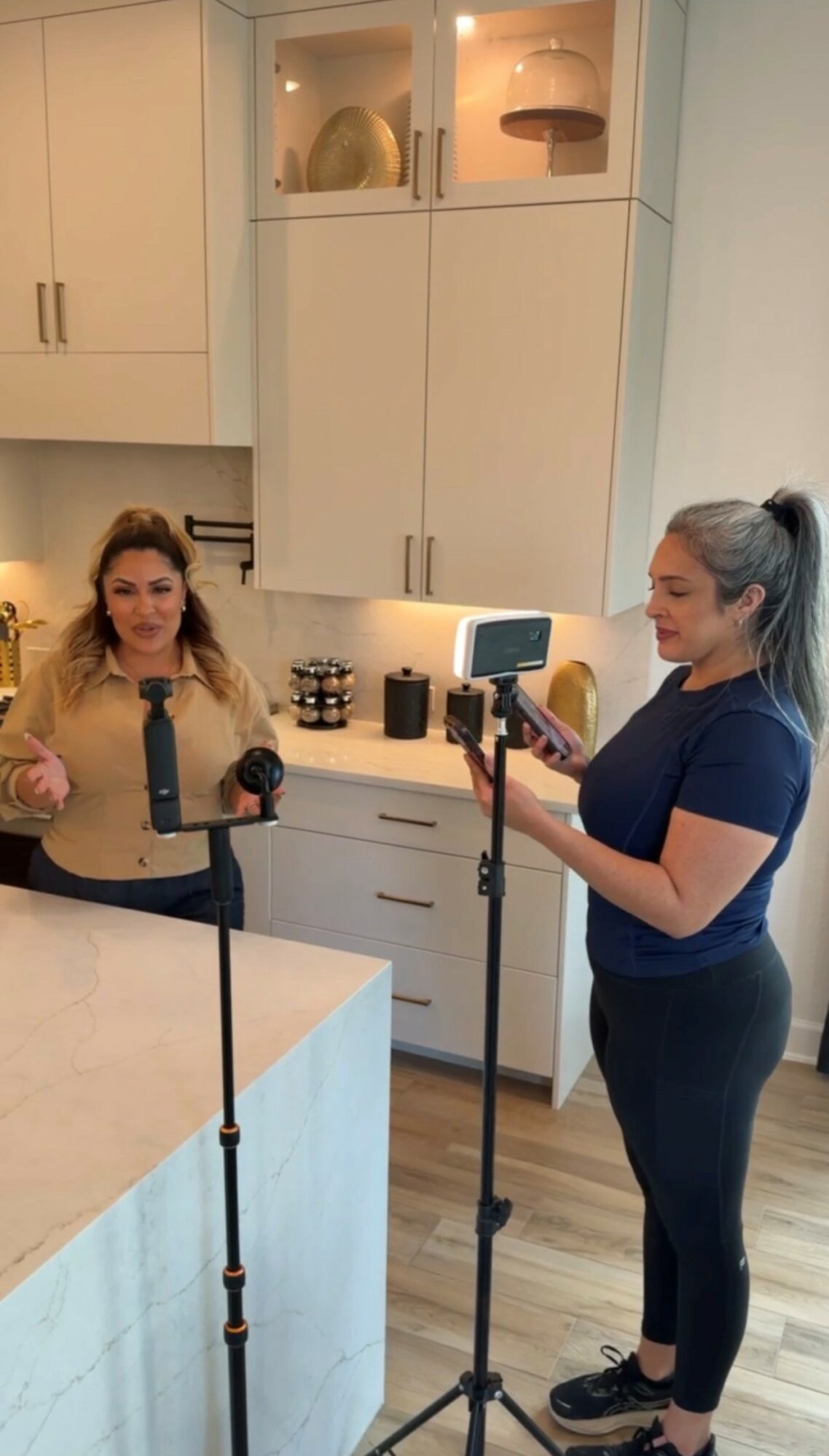 Two women in a kitchen, one taking a photo with a smartphone, the other standing near a countertop, smiling.