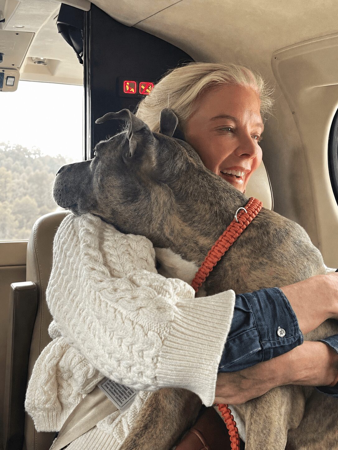 Woman smiling with a large dog on her lap inside an airplane, window view outside, woman wearing a white sweater.