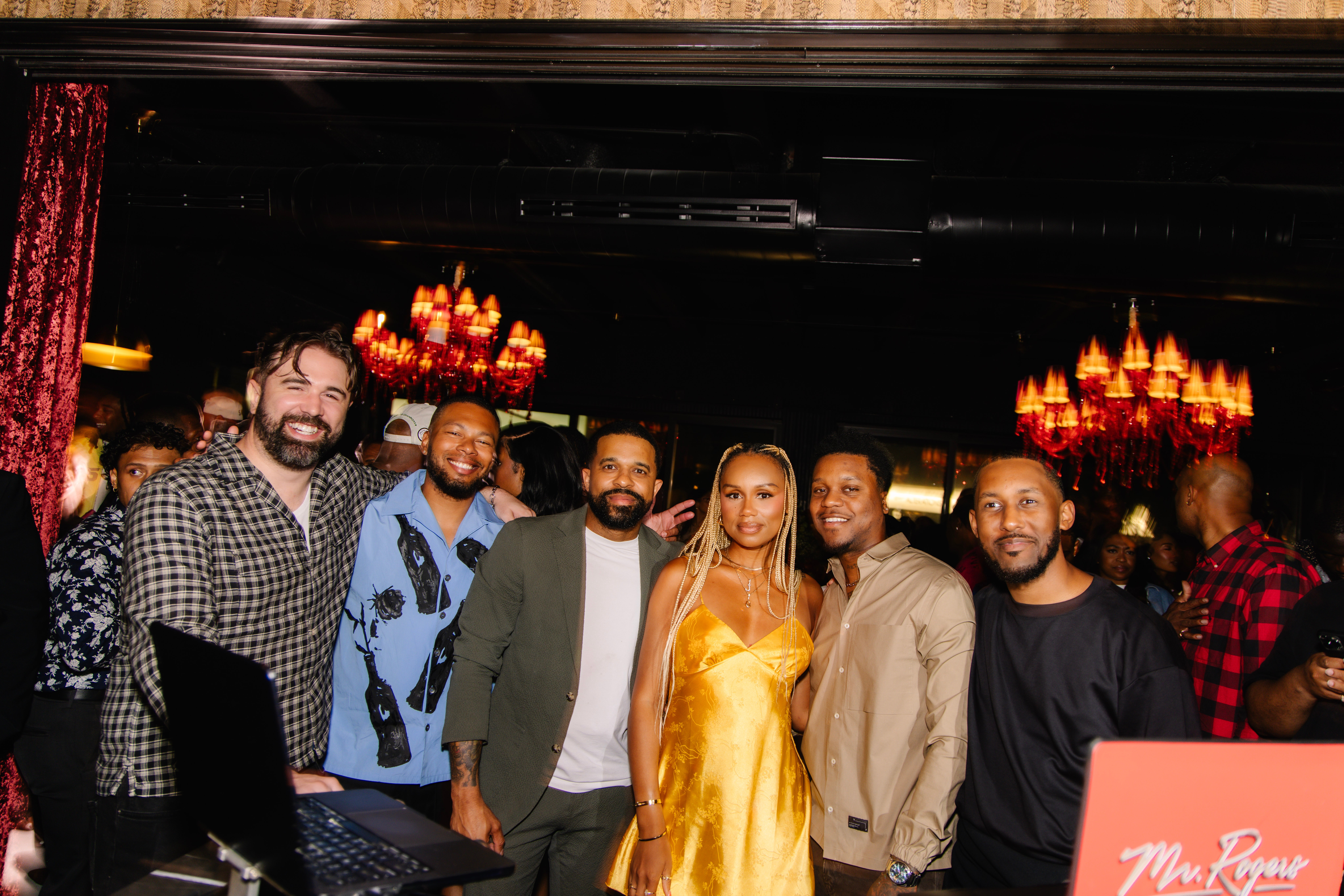 Group of six people posing together in a dimly lit venue with chandeliers, smiling at the camera.