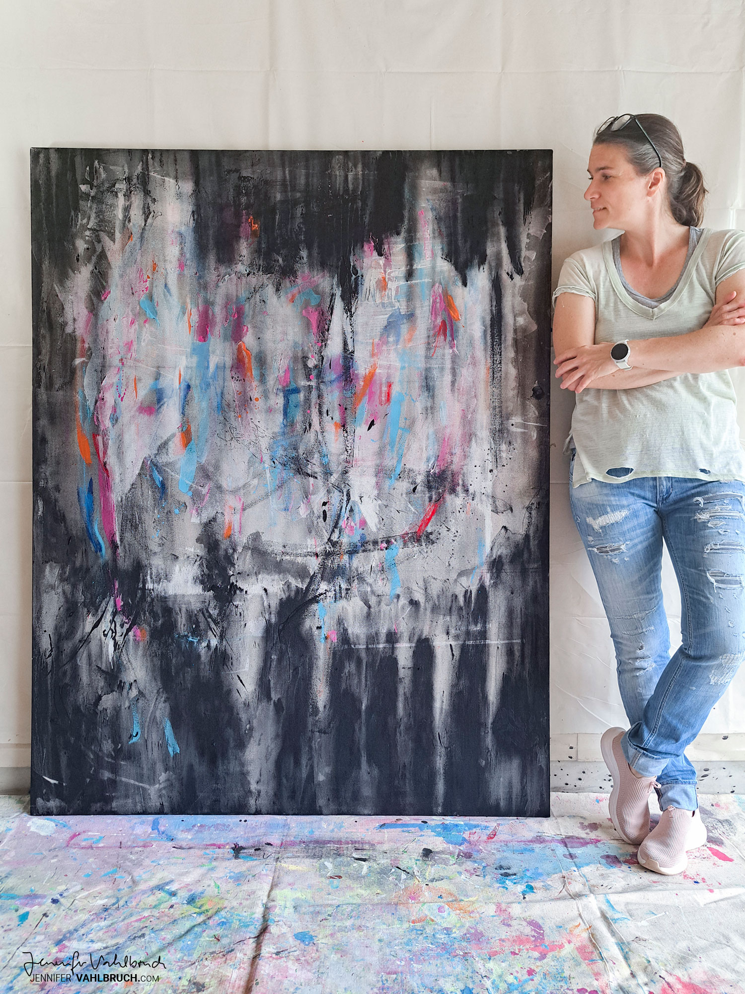 Woman standing next to an abstract painting on a canvas, with a colorful paint-splattered floor beneath her.