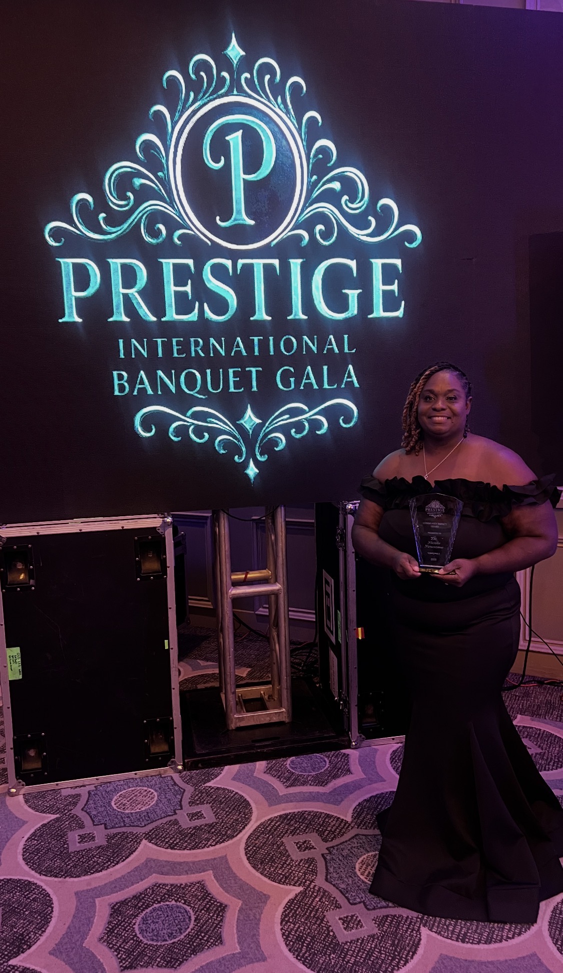 Woman in black dress holding an award at a gala, with a large illuminated logo behind her, on a patterned carpet.