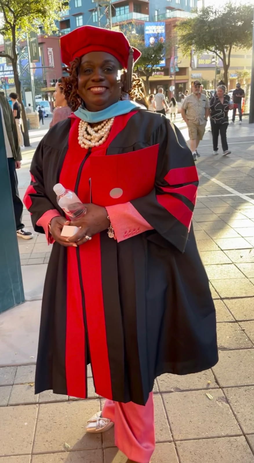 Person in graduation gown and cap smiling outdoors, holding a diploma, with city street background.