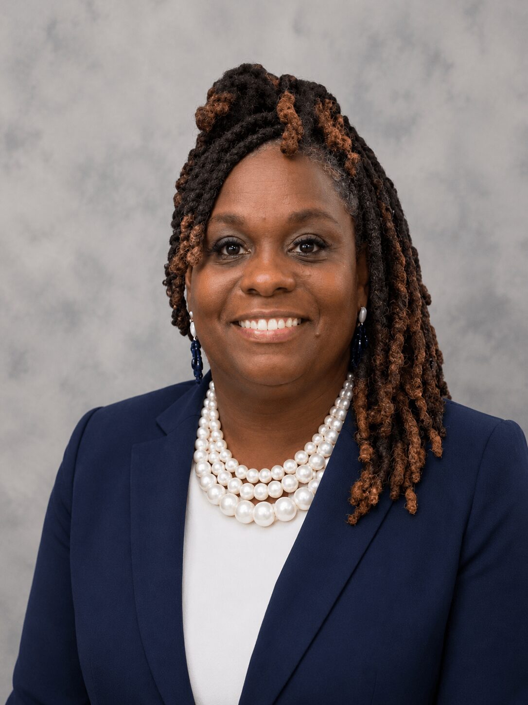 Portrait of a woman with styled dreadlocks, wearing a navy blazer and pearl necklace, smiling against a light gray background.
