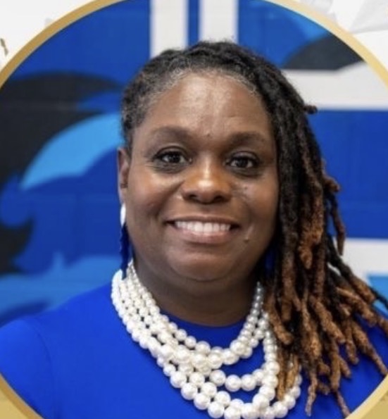 Woman with braided hair wearing a blue top and pearl necklace, smiling, with a blue and black background.